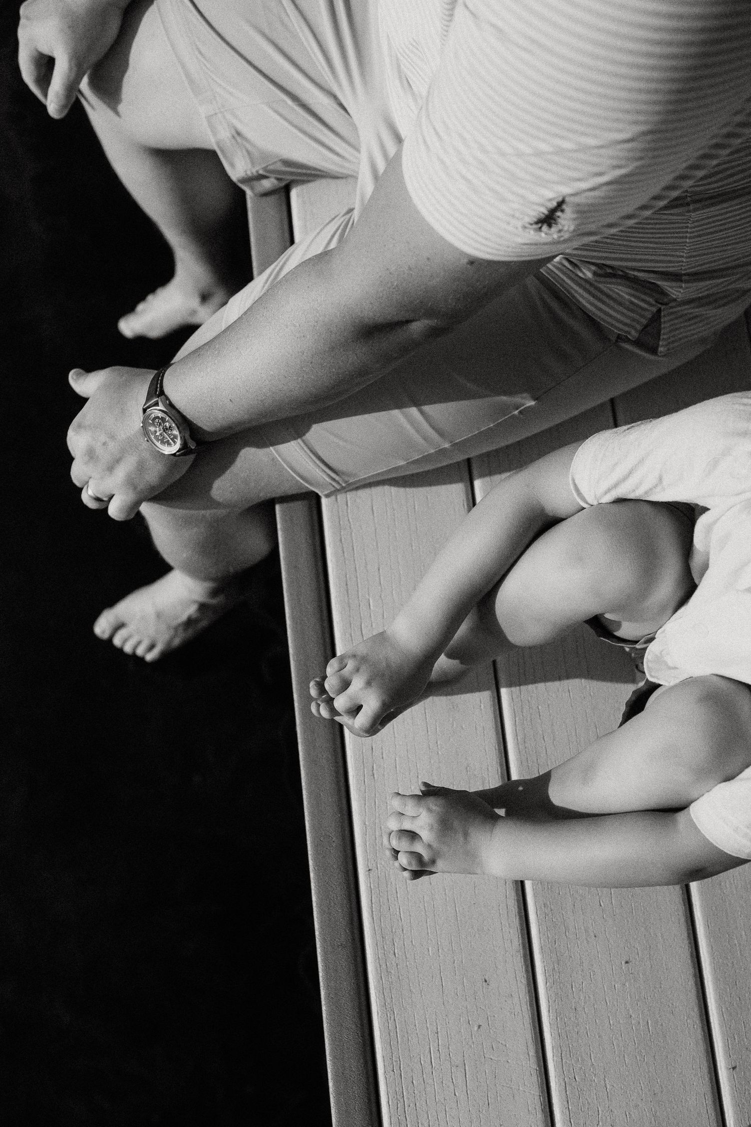 Black and white overhead view of family sitting on wooden dock with feet dangling over water.