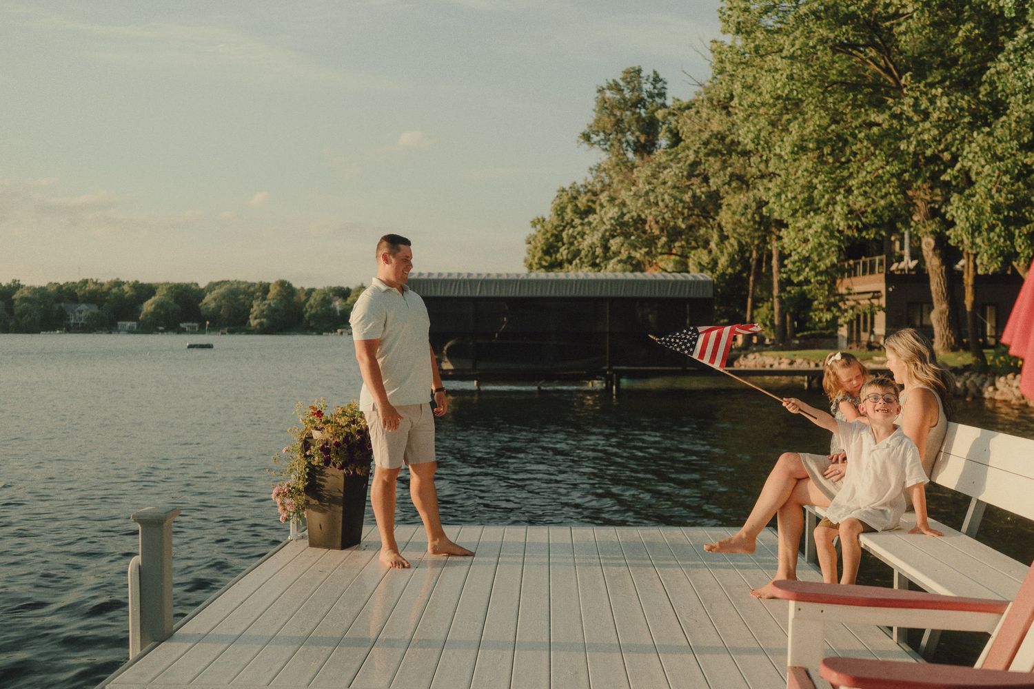 Family relaxing on white dock by lake with boathouse at golden hour.