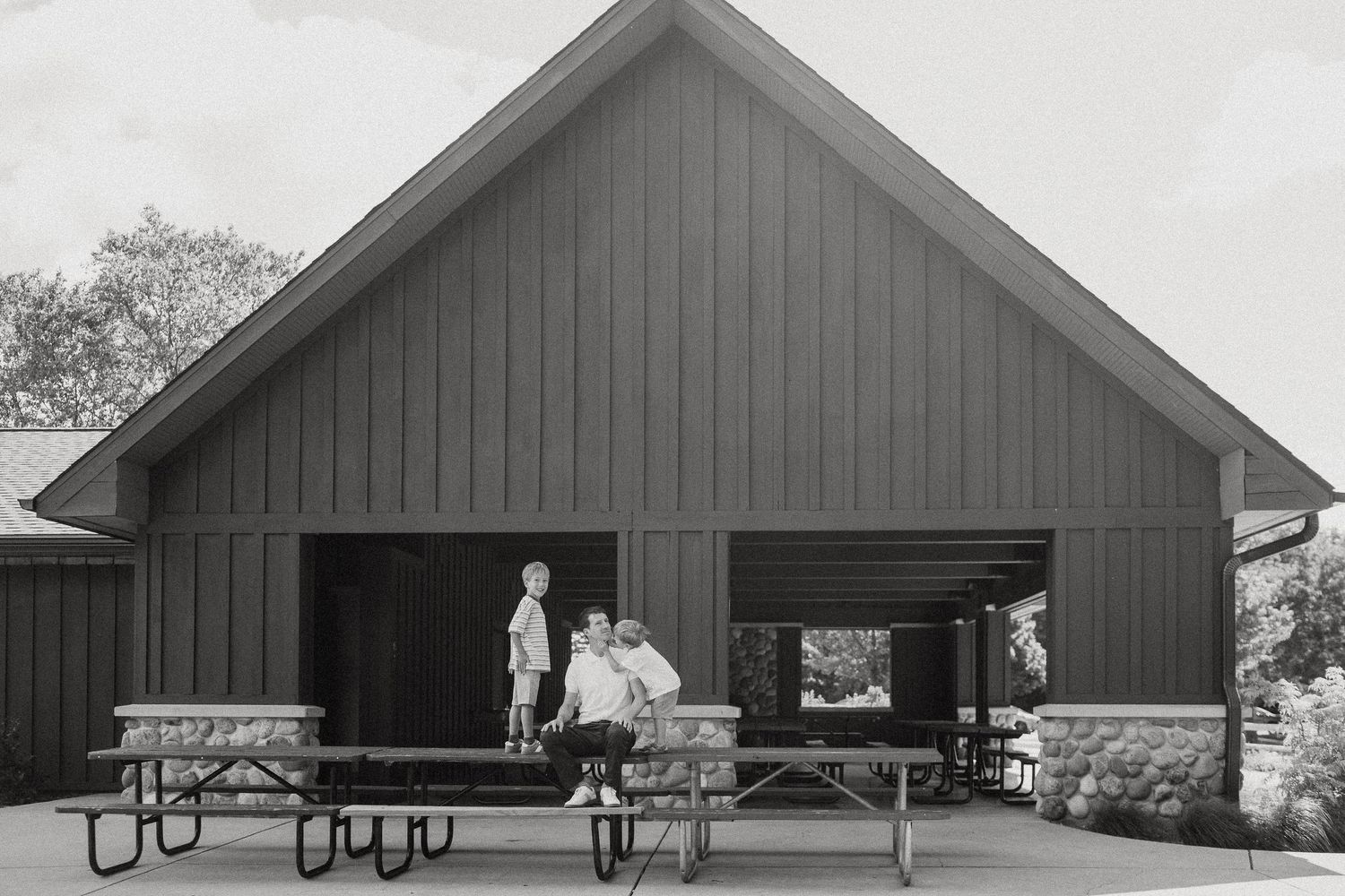 Black and white photo of a father and son relaxing on picnic tables outside a barn pavilion.