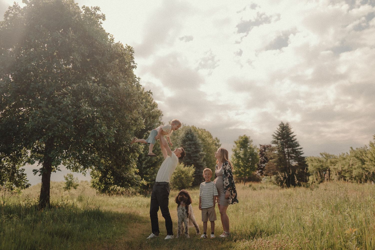 Family of five enjoying a sunny day in a lush green meadow, with father lifting toddler in the air.