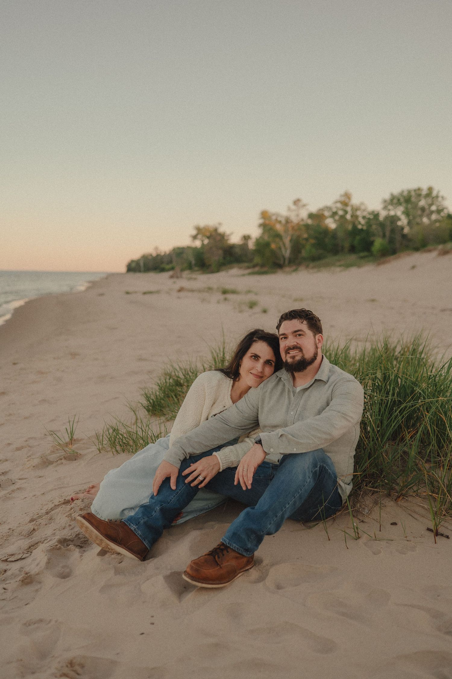 Couple sits together on sandy beach dunes at sunset with ocean in background.