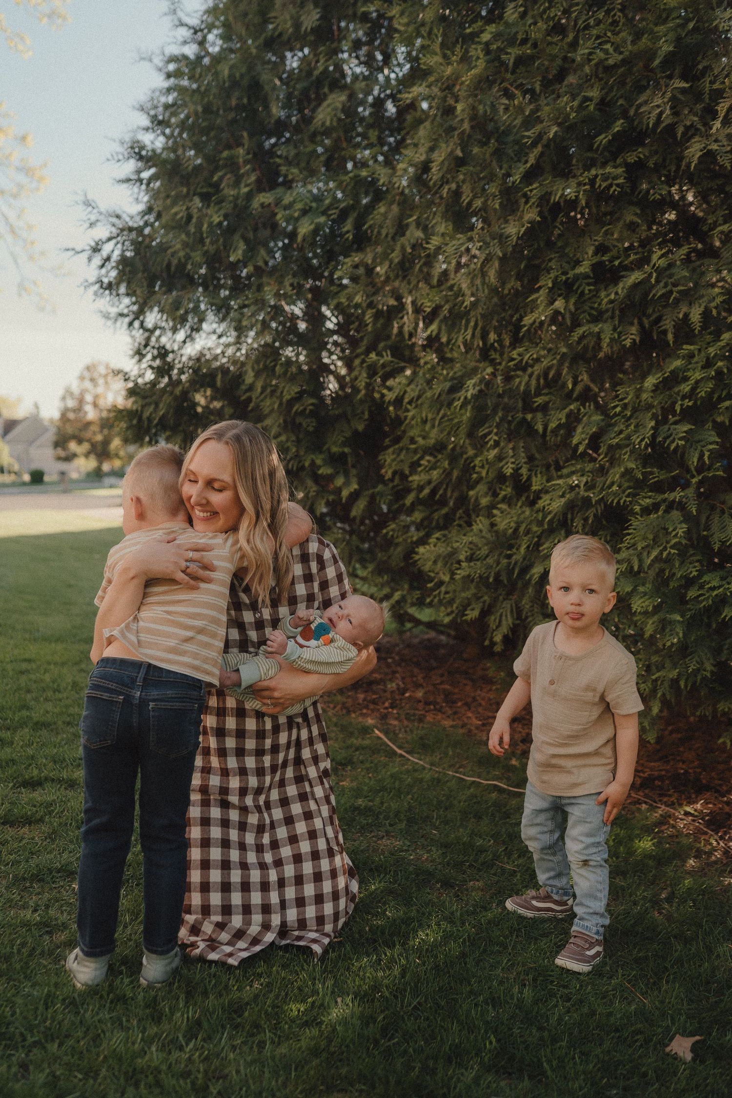 Smiling blonde mother in plaid dress holding two children near evergreen tree in sunny backyard.