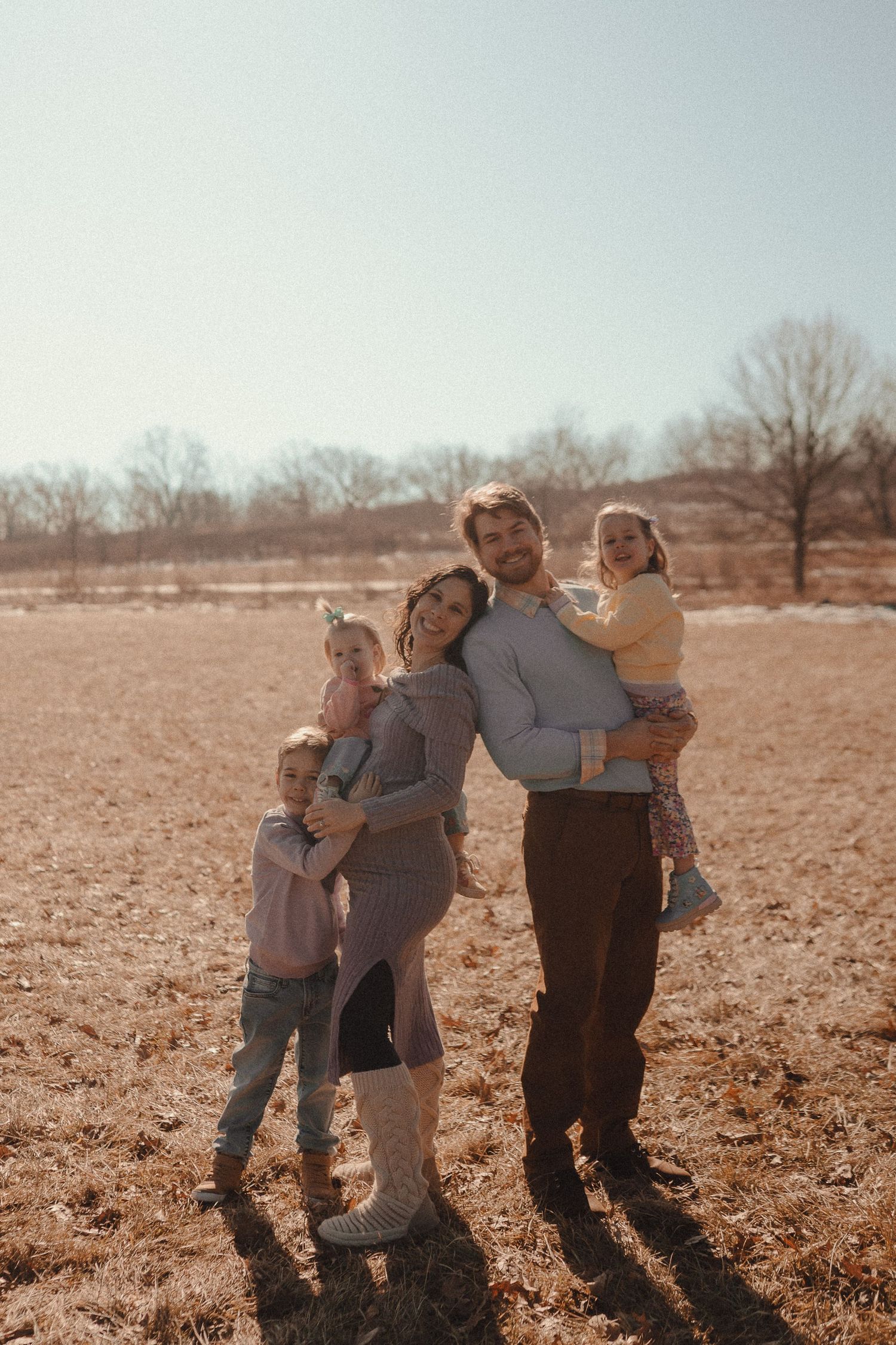 Family members embrace while standing in a barren field on a sunny afternoon.