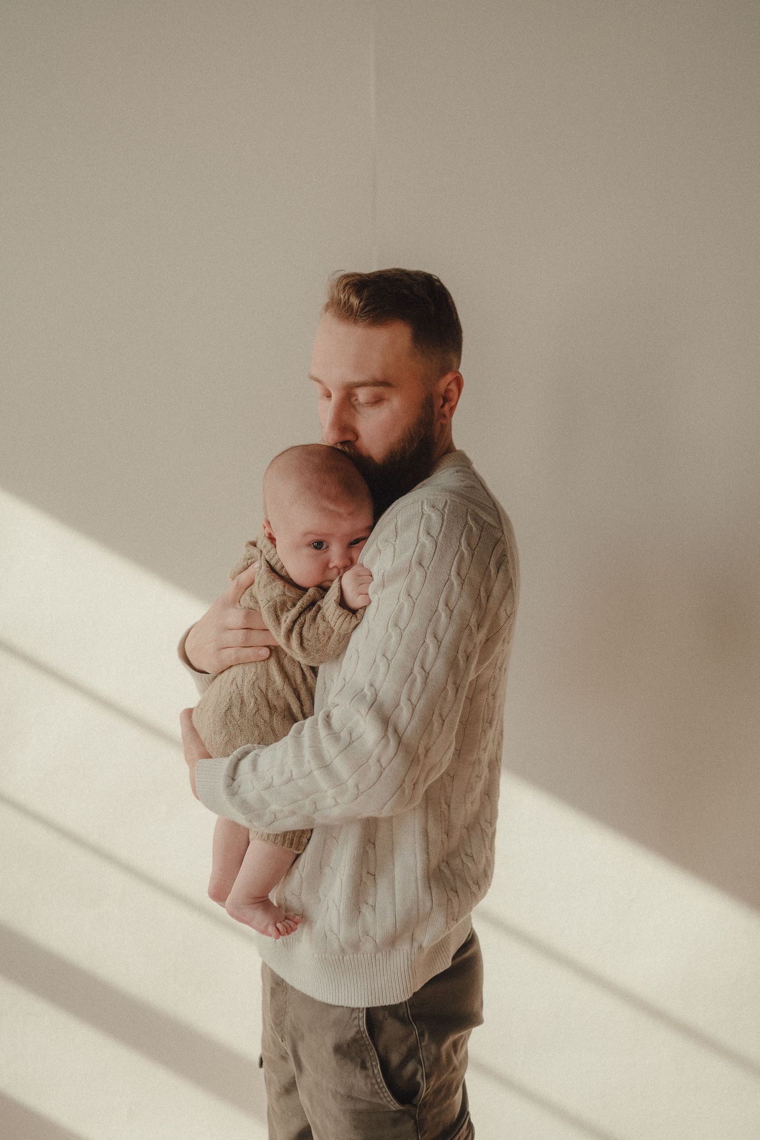 A tender moment captured as a person in a cream cable knit sweater holds a baby in matching neutral tones against a sunlit wall.