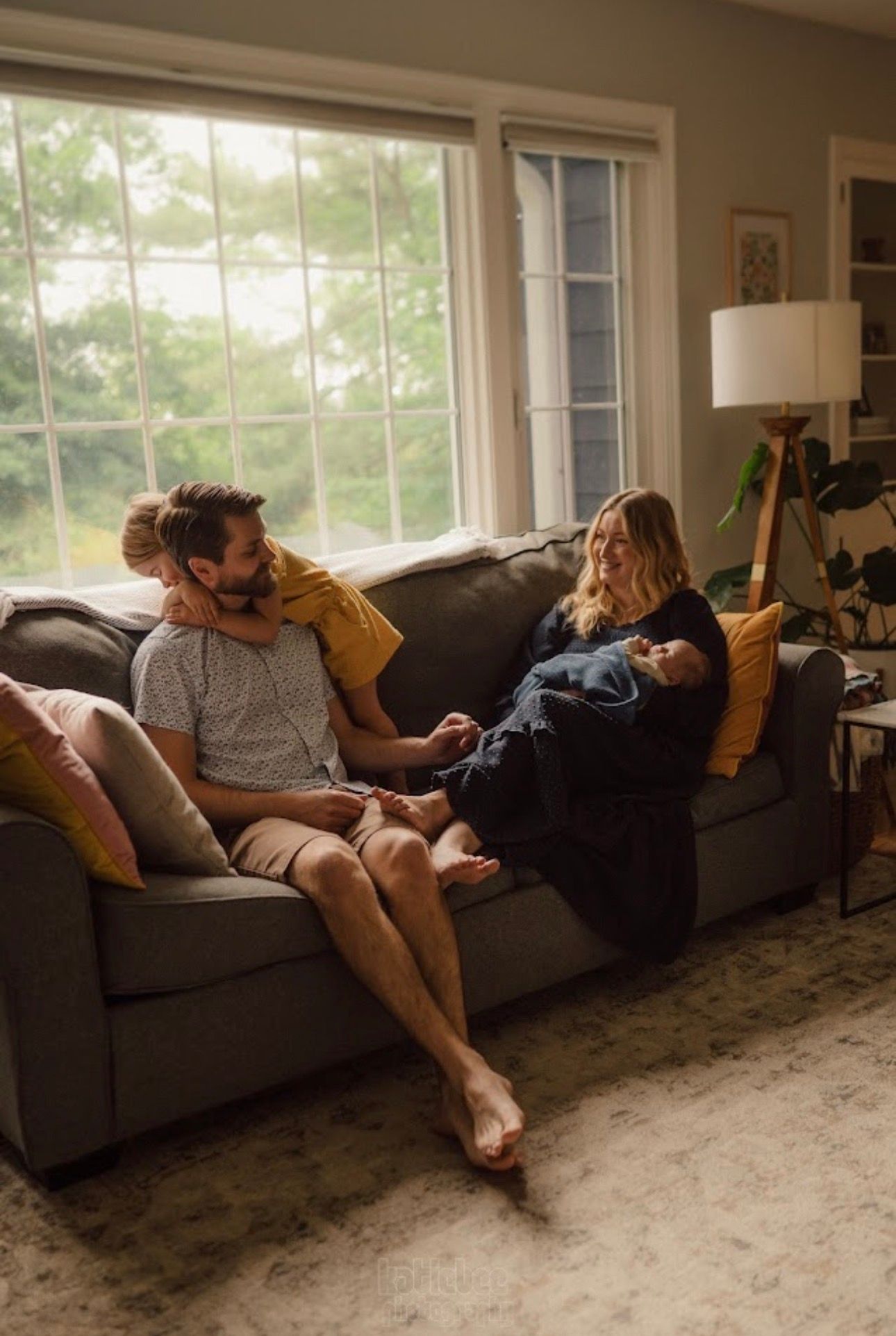 A cozy scene of people lounging together on a grey sofa in a sunlit living room.