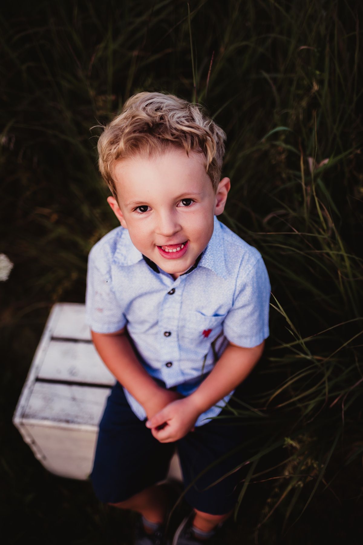 A young child in blue shirt sits on a wooden crate outdoors smiling at the camera.