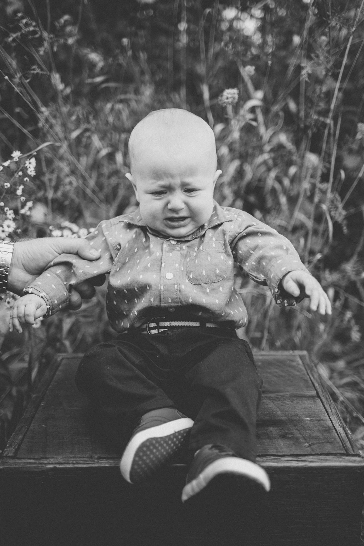 Black and white image of crying baby sitting on dark surface outdoors.