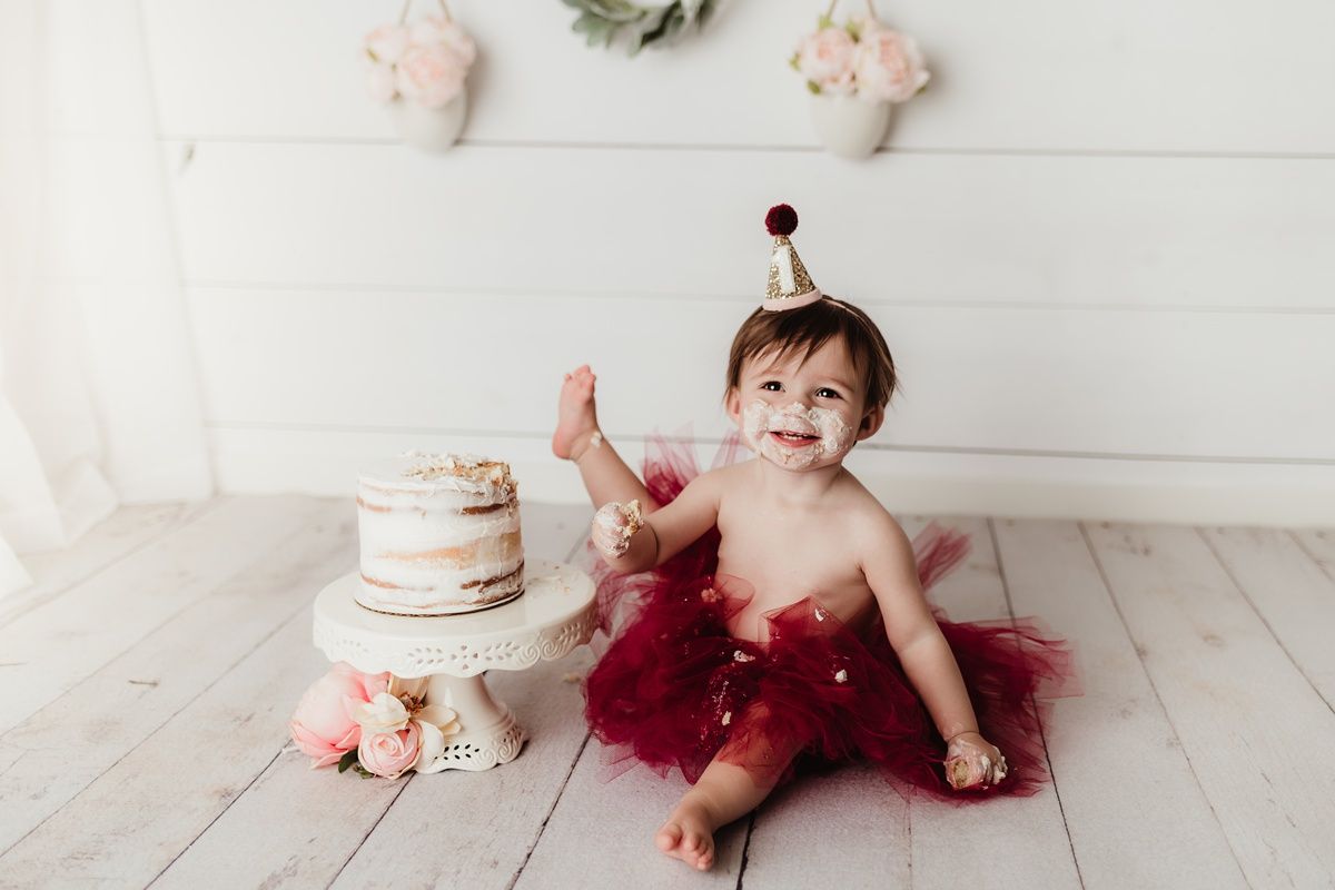 Baby in burgundy tutu sits next to first birthday cake on white wooden floor.