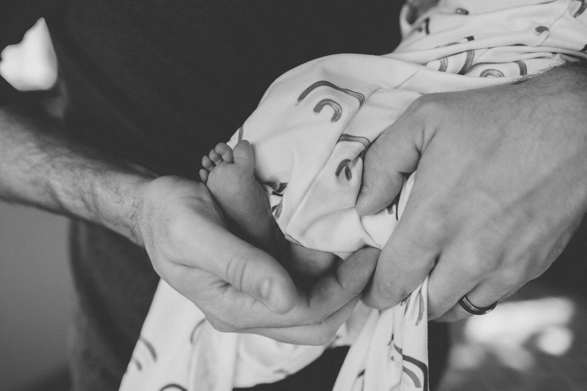 Black and white close-up of gentle hands cradling newborn in patterned outfit.