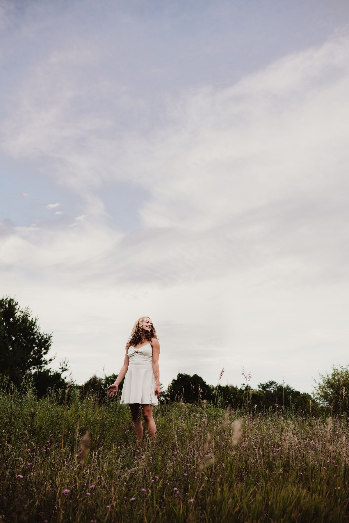 Figure in white dress standing in tall grass field under cloudy summer sky.