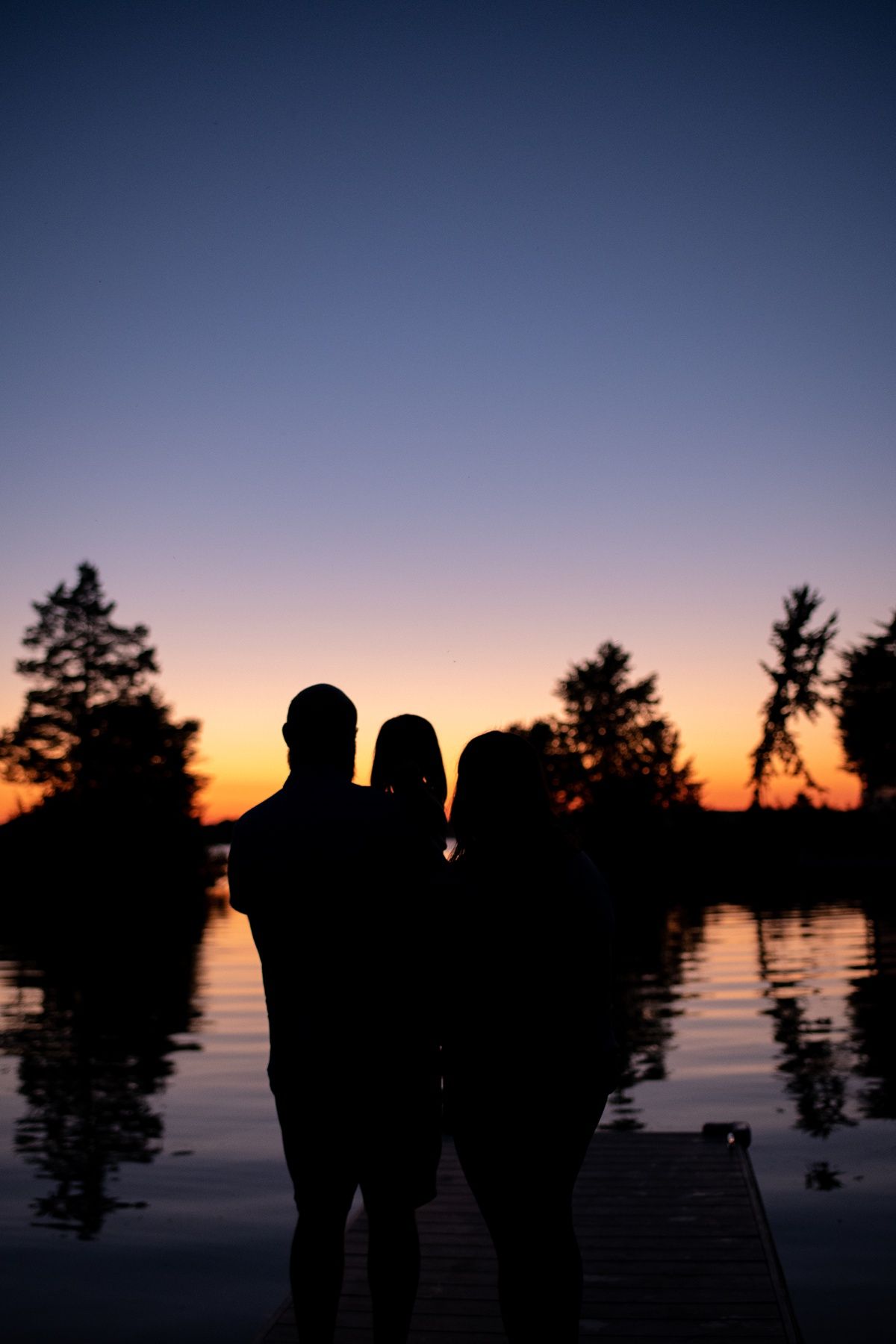 Silhouettes of people standing on a dock during sunset with trees reflecting on calm water.
