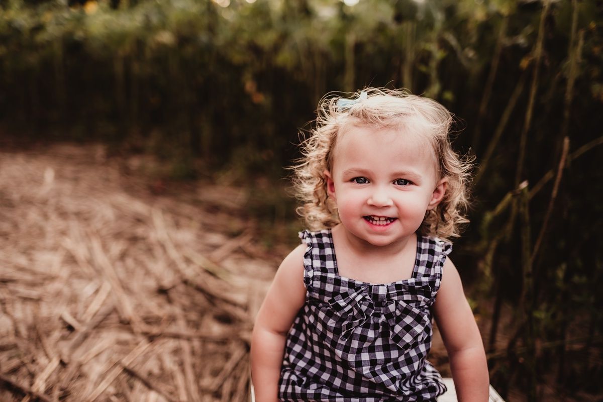 Child wearing gingham dress smiling in sunflower garden.