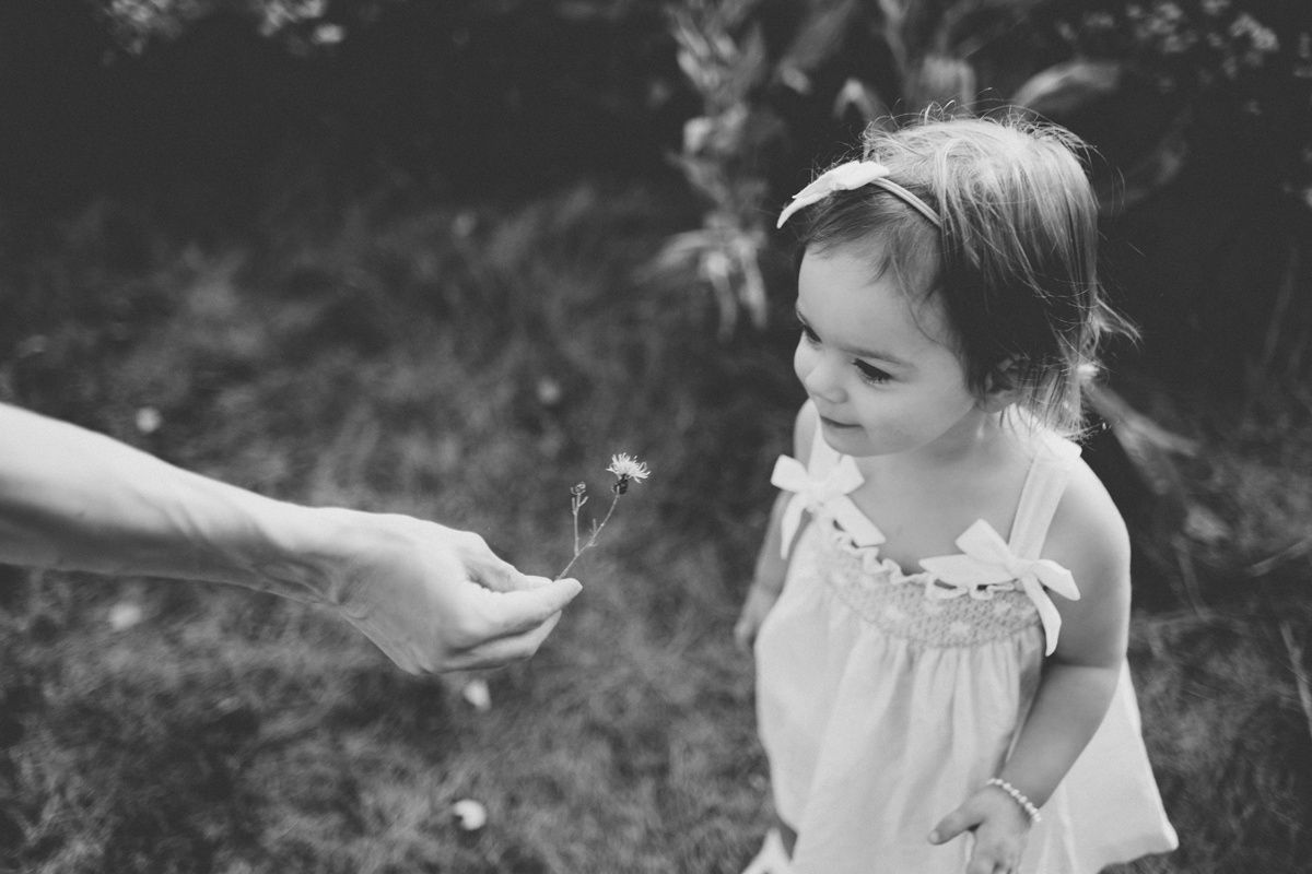 A black and white photo of a young child in a white dress reaching towards a dandelion being offered.