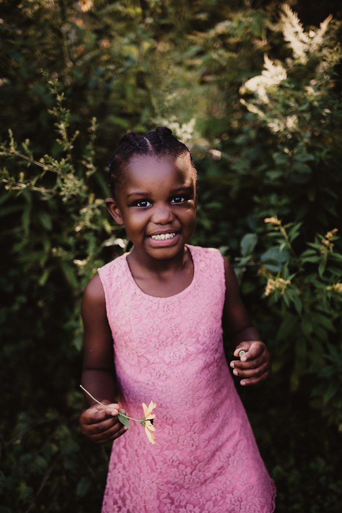 A bright smile lights up the scene as someone holds a flower in a pink dress outdoors.