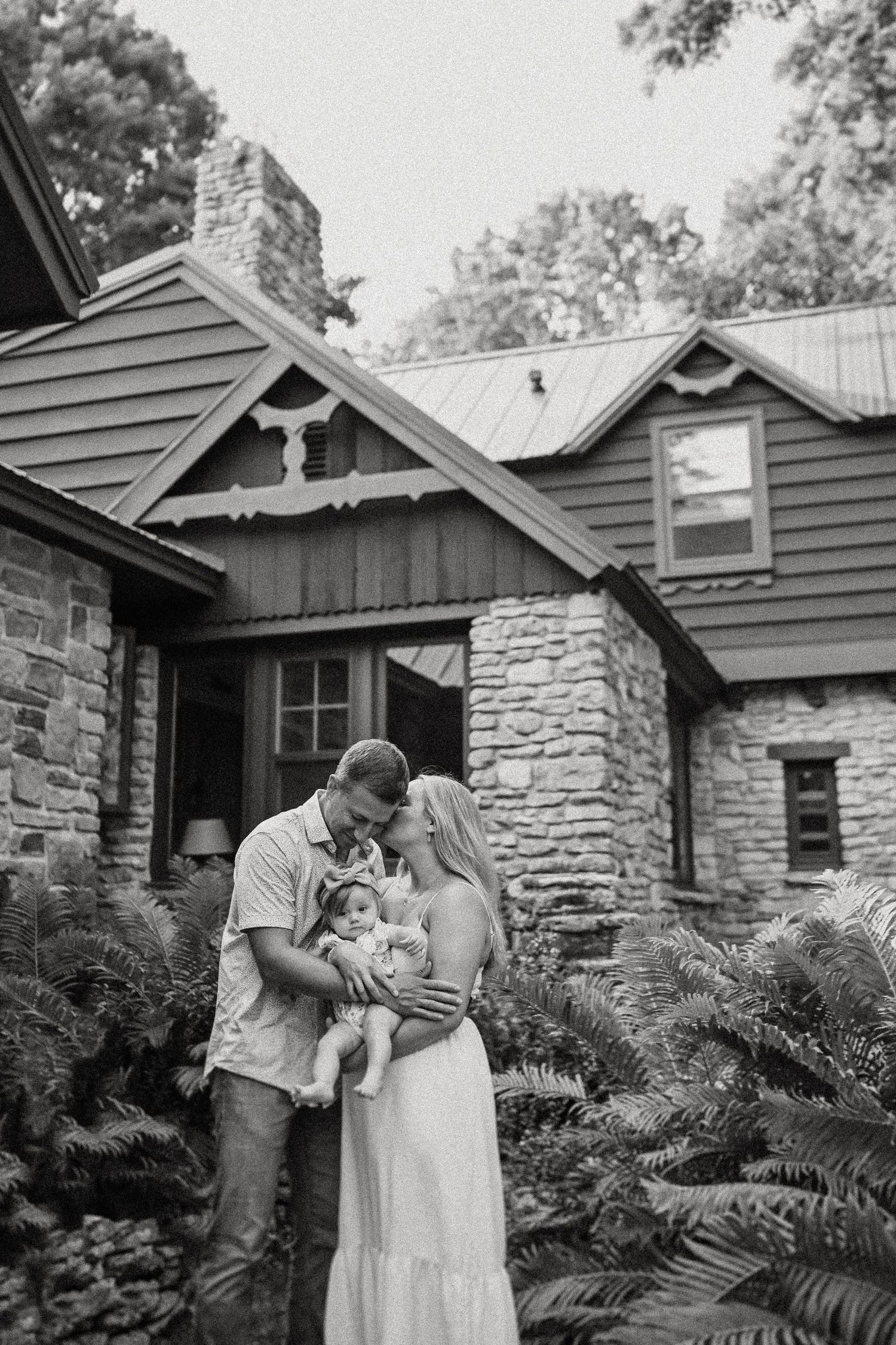 A couple holds their baby outside a rustic stone and wood cottage surrounded by ferns.