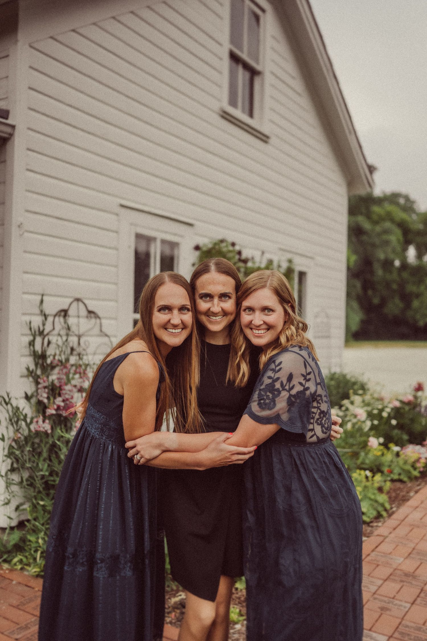 Three women in navy dresses embrace in front of a white house with pink flowers growing nearby.