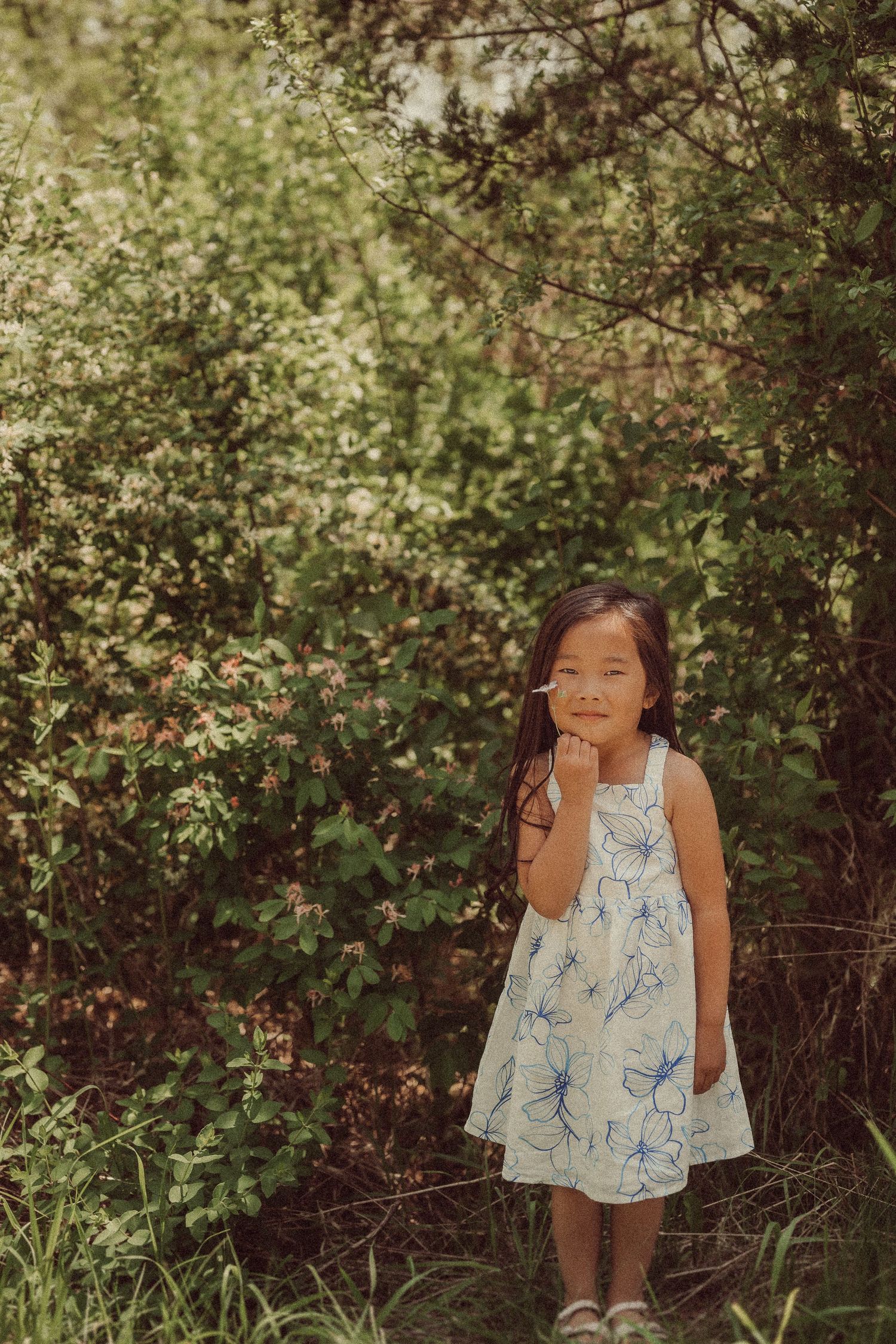 A floral dress-clad figure poses thoughtfully against a backdrop of green bushes and trees.
