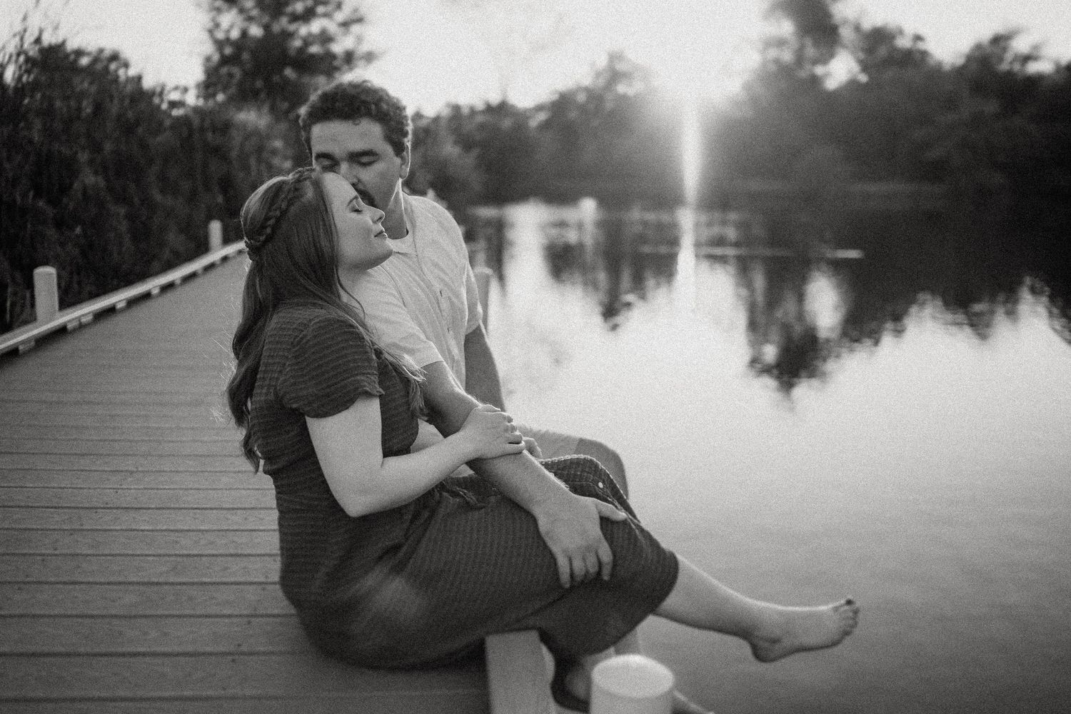 A couple sits close together on a wooden dock at sunset over a calm lake.