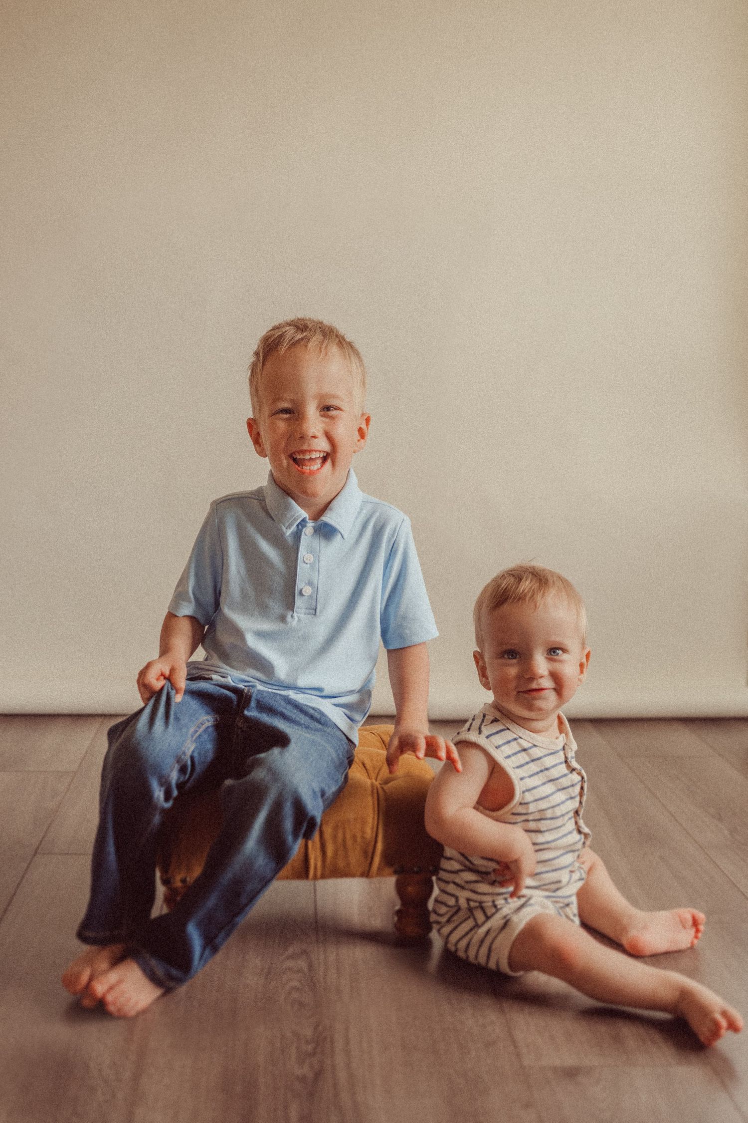 Two children sitting together on a wooden bench against a neutral wall with hardwood flooring.