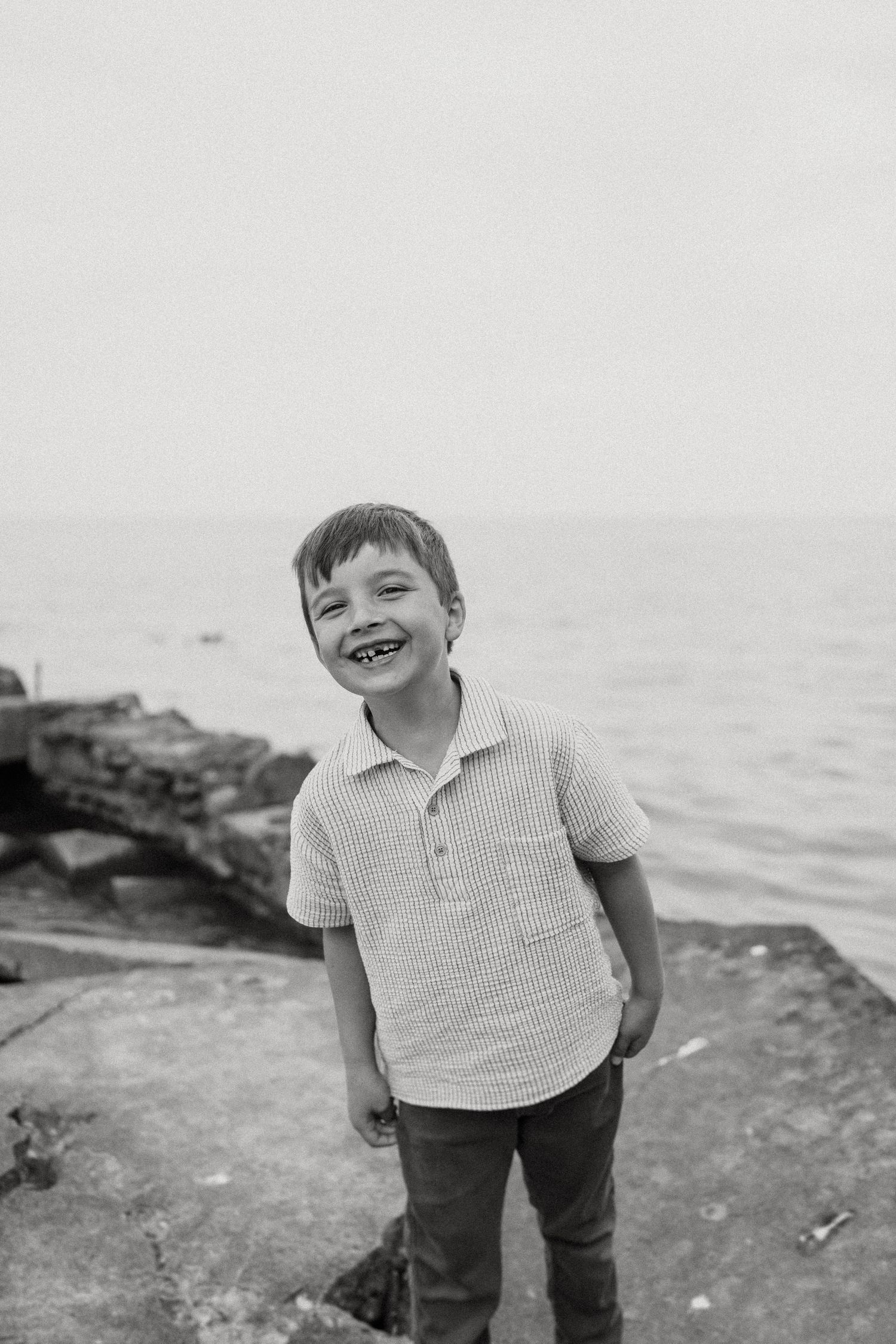Black and white portrait captures a bright smile on the rocky beach with ocean waves in background.