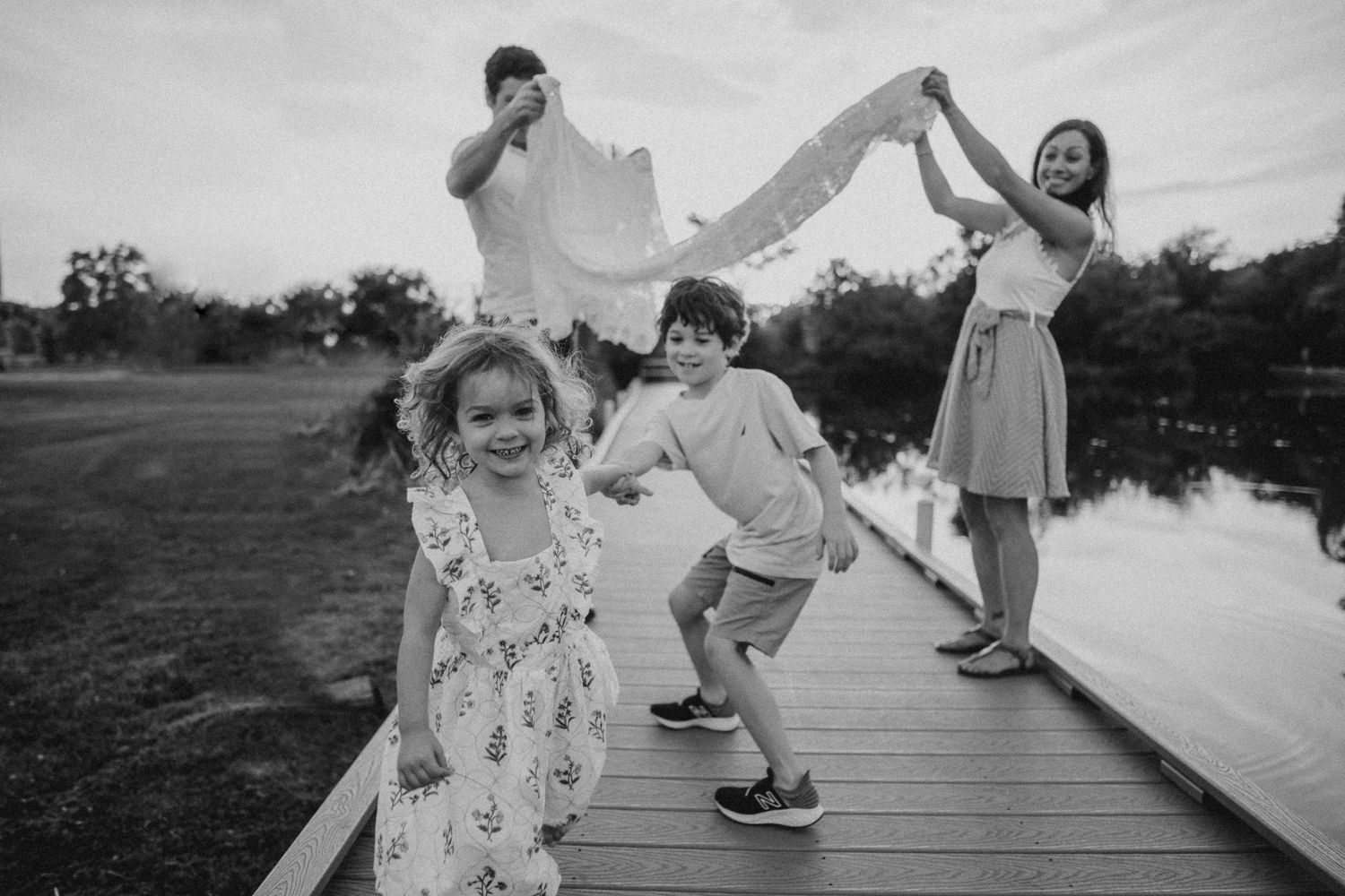 A joyful family plays and dances on a wooden dock over water in black and white.