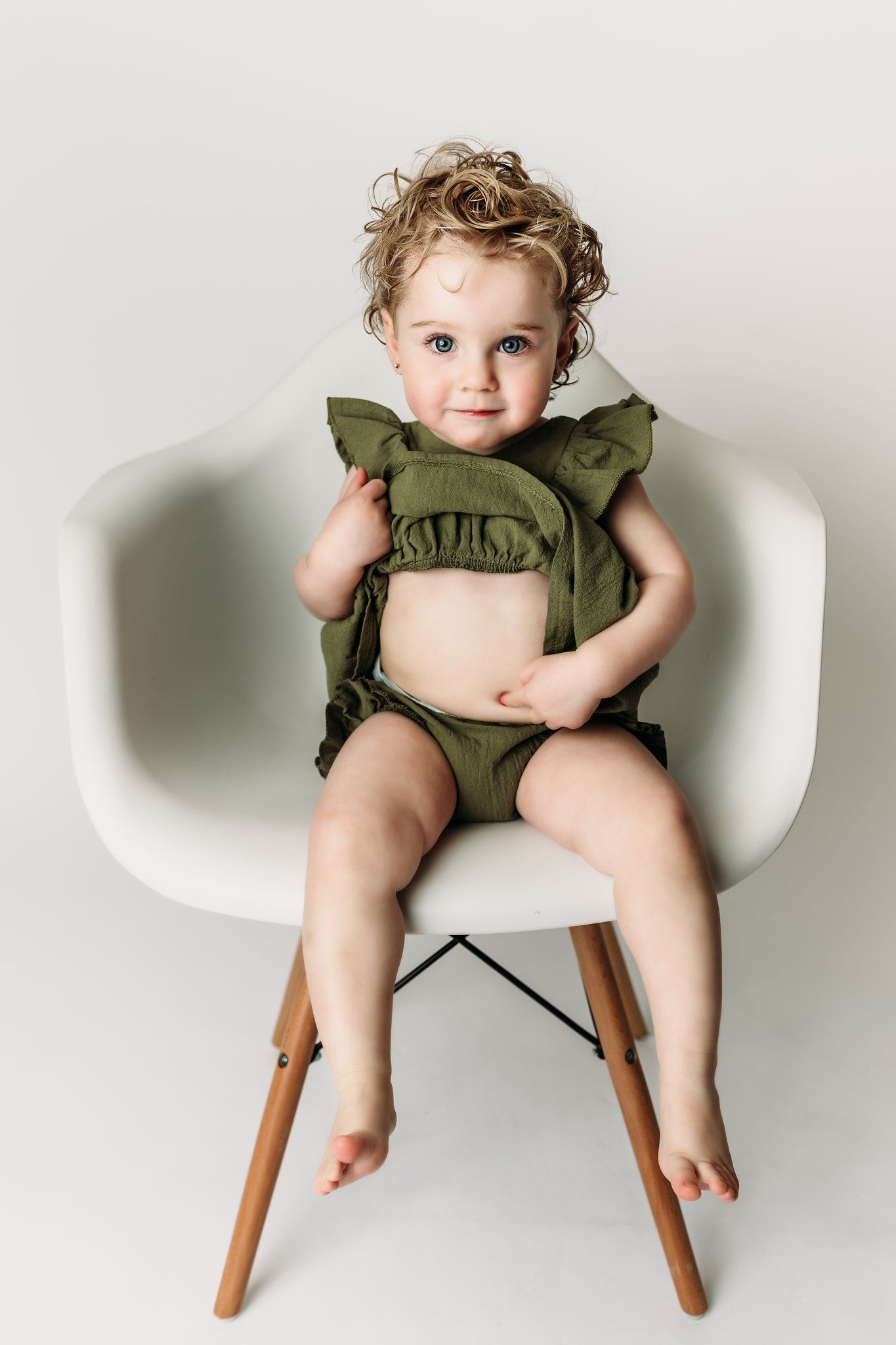A small child sits in a modern white chair wearing an olive green outfit with ruffles.
