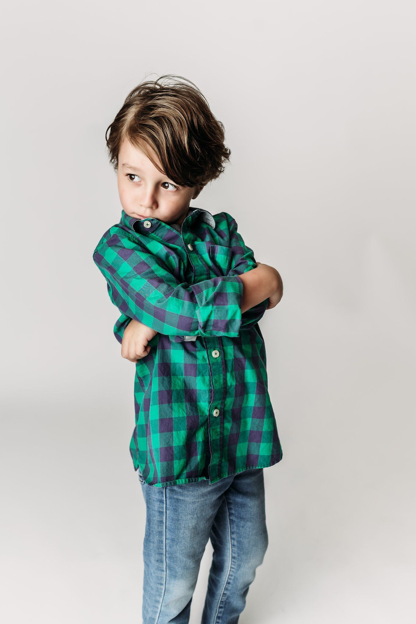 Child in green plaid shirt stands with crossed arms and serious expression against white background.