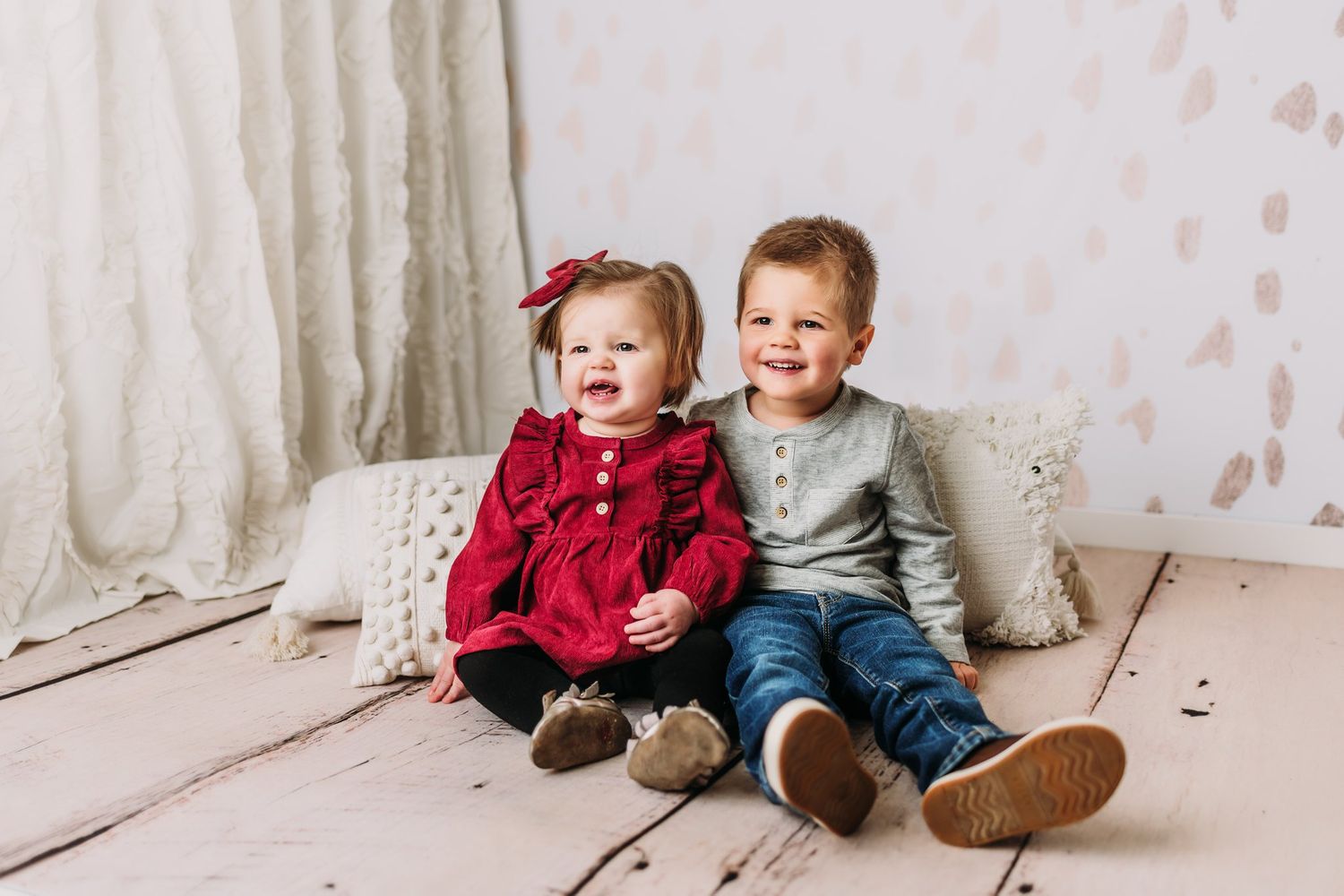 Two young children sitting together on white wooden floor with textured backdrop.