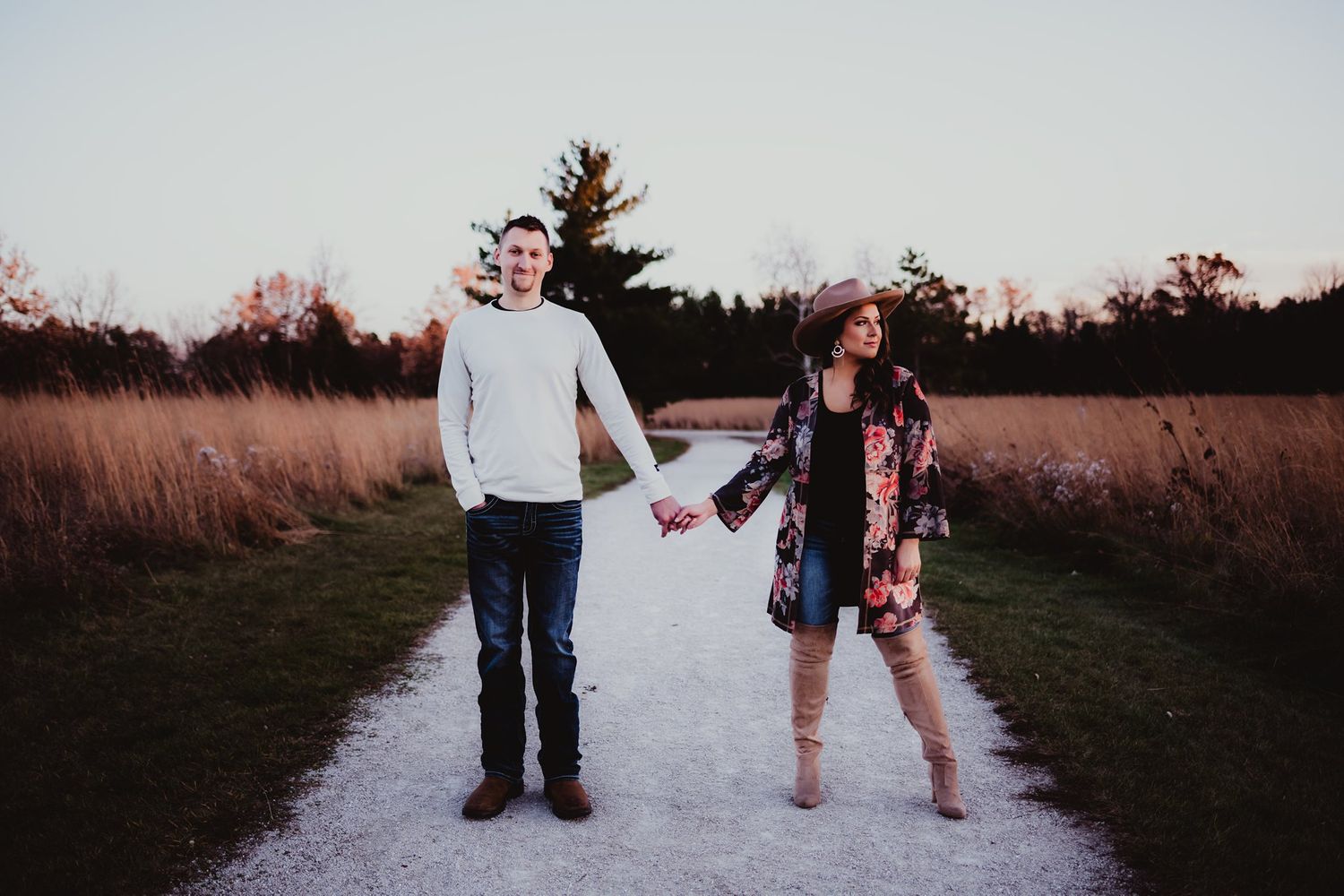 A couple walks hand in hand on a path through golden grass at sunset.