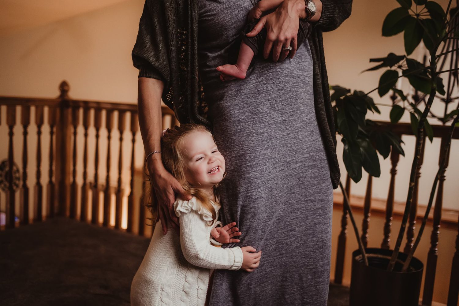 A tender embrace showing a child hugging someone wearing a gray dress near a wooden banister and houseplant.
