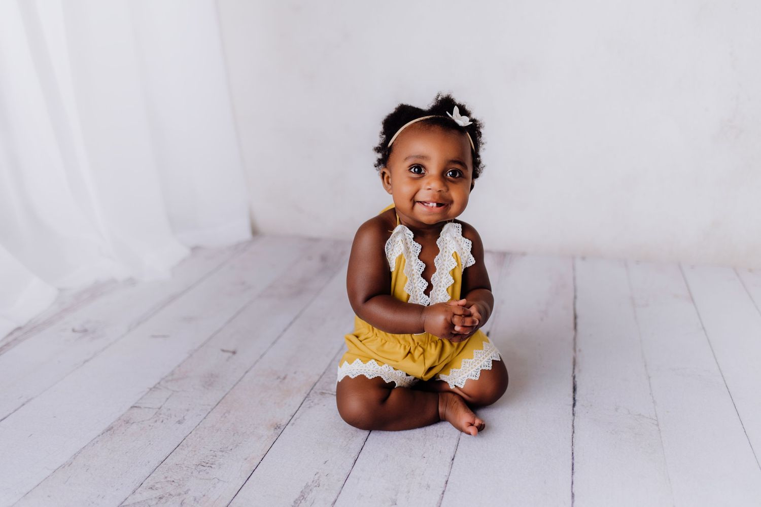 Joyful baby in yellow outfit sitting on white wooden floor during photo session.