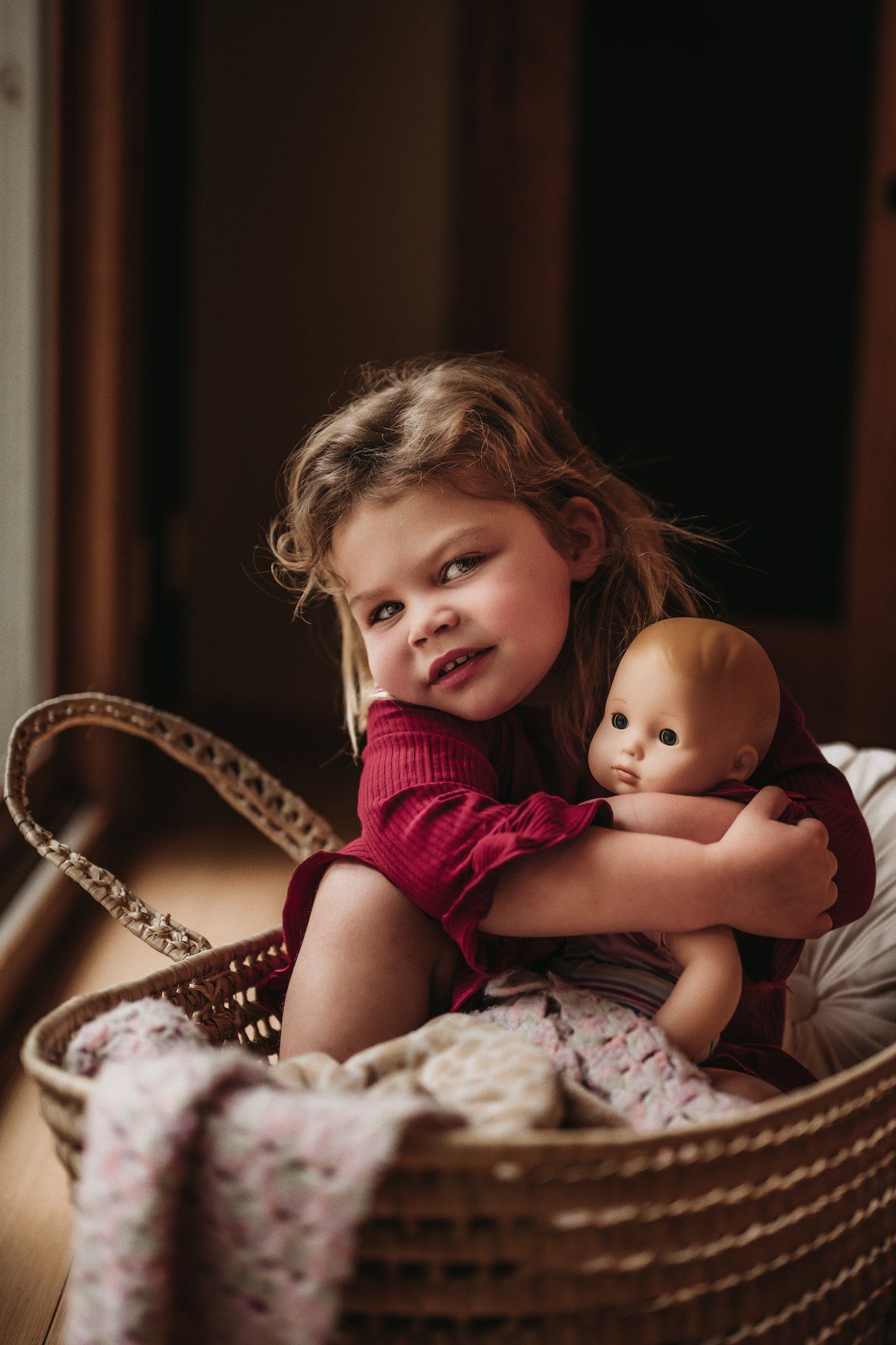 Young child in red sweater sits in woven basket holding baby doll.