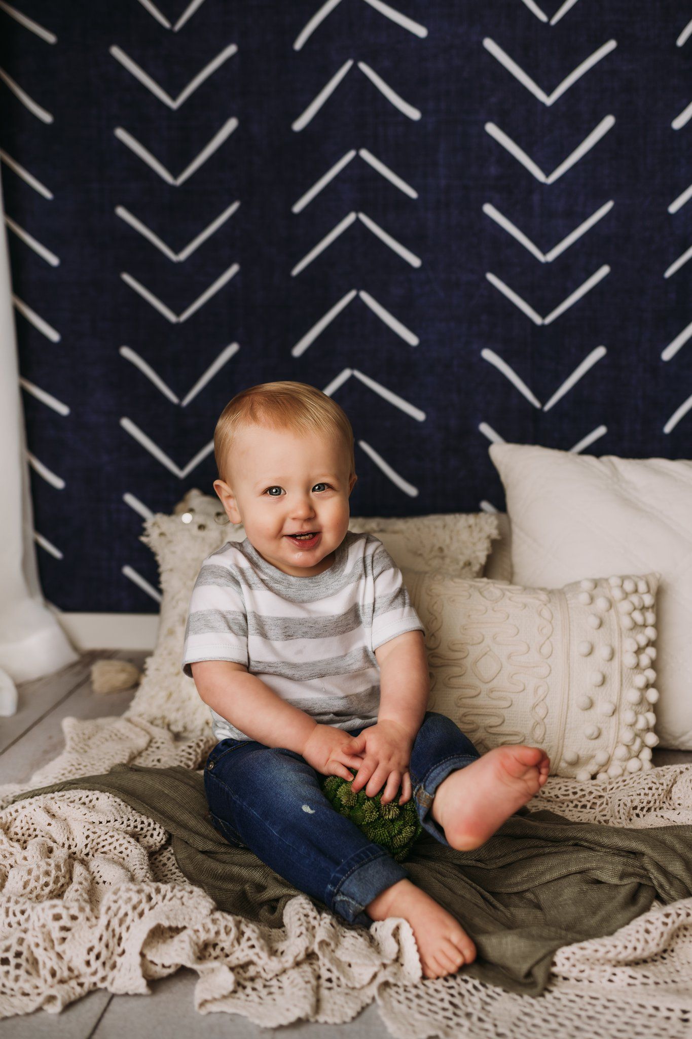 A small child in stripes sits among cozy textured blankets against a geometric backdrop.