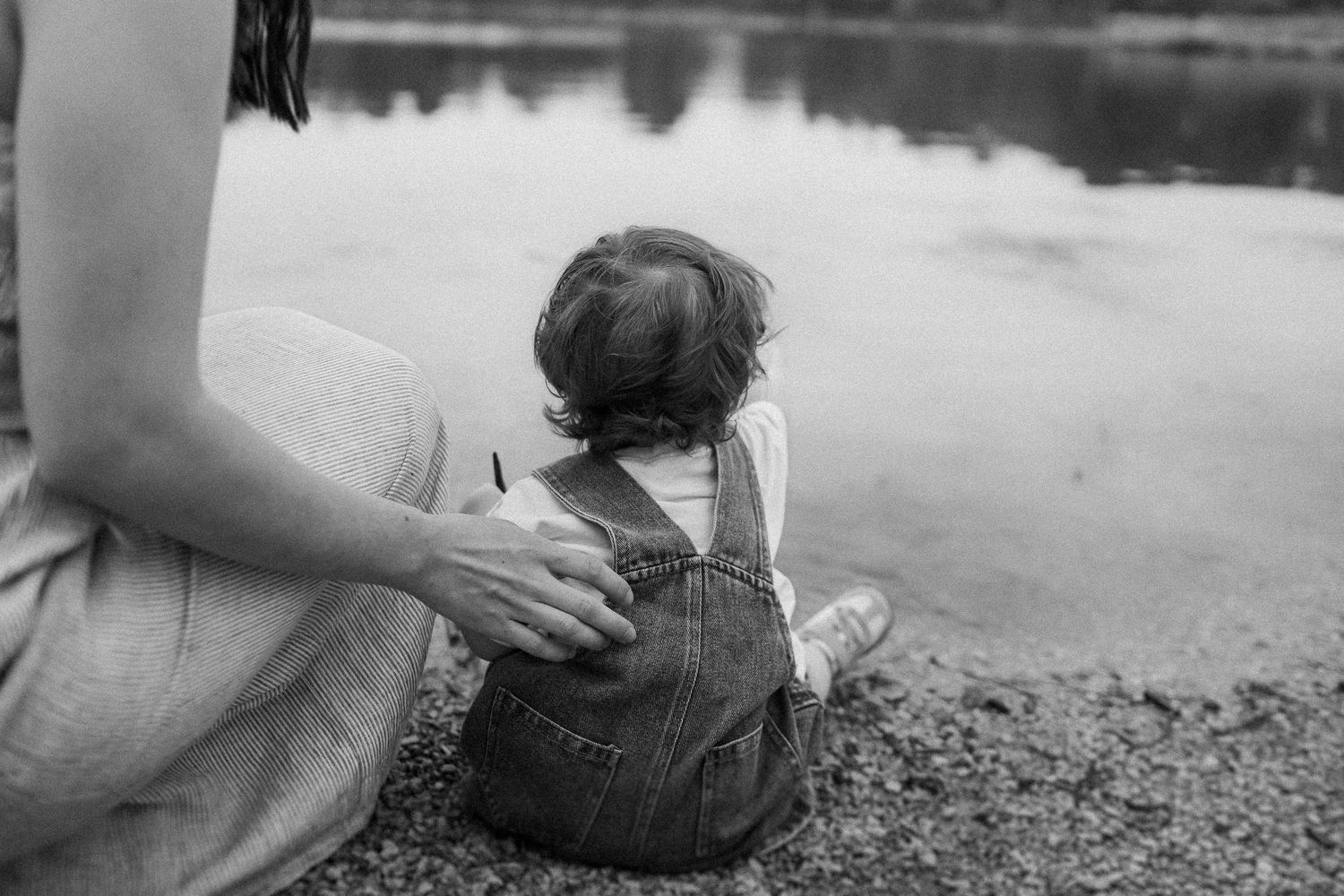 A quiet moment by the lake as a child in denim overalls sits at the water's edge with an adult.