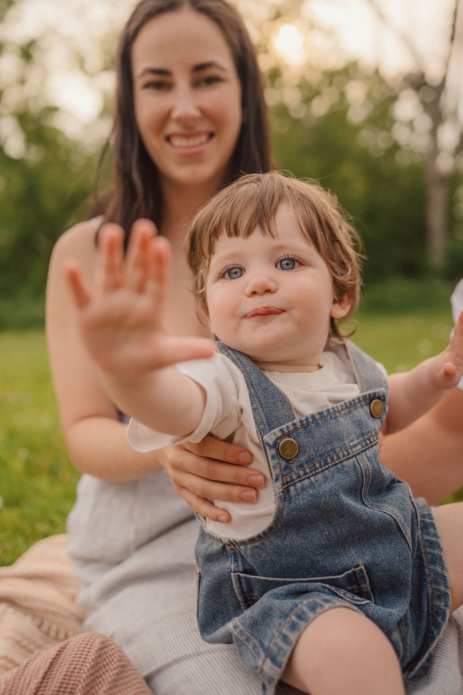 Child in denim overalls sits outdoors with parent on sunny day, reaching towards camera with a happy expression.