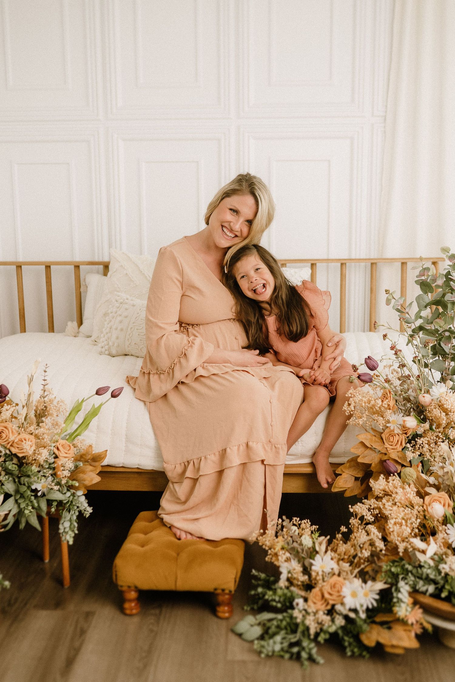 Two people in matching blush pink dresses sit together on a wooden daybed surrounded by dried floral arrangements.