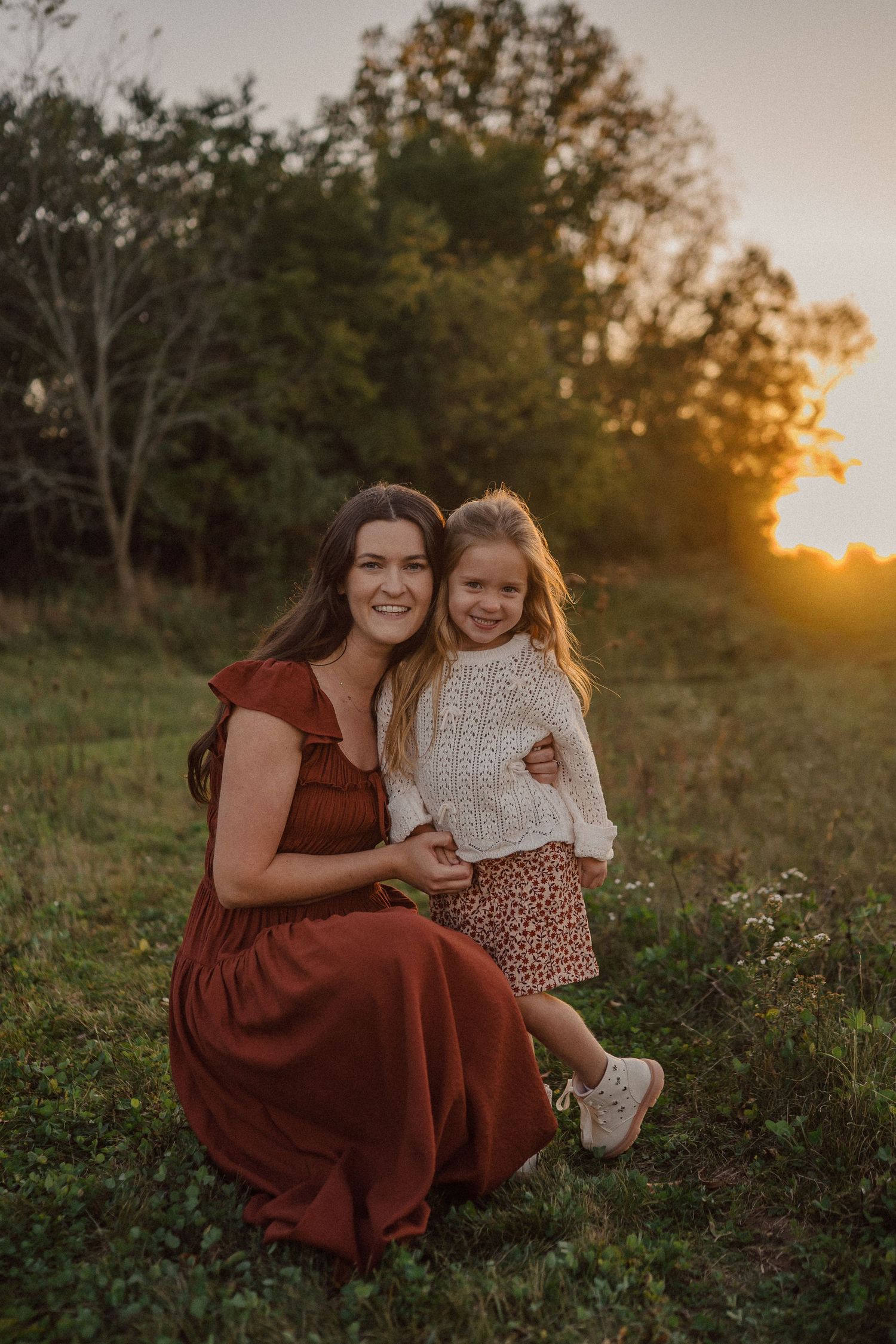 A mother and daughter share a tender moment in a field during golden hour sunset.