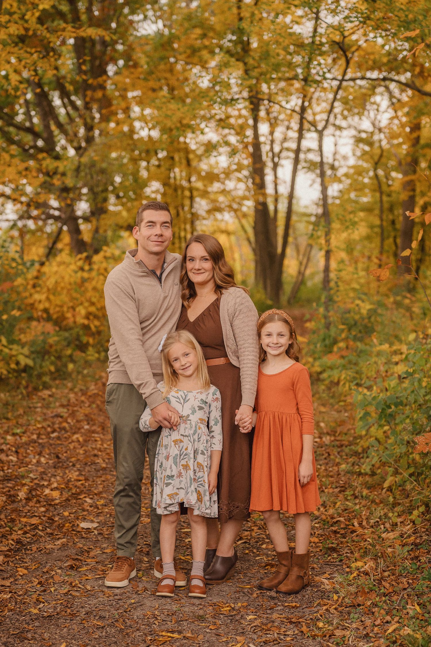 A family poses together in an autumn forest setting with fallen leaves and golden foliage in the background.