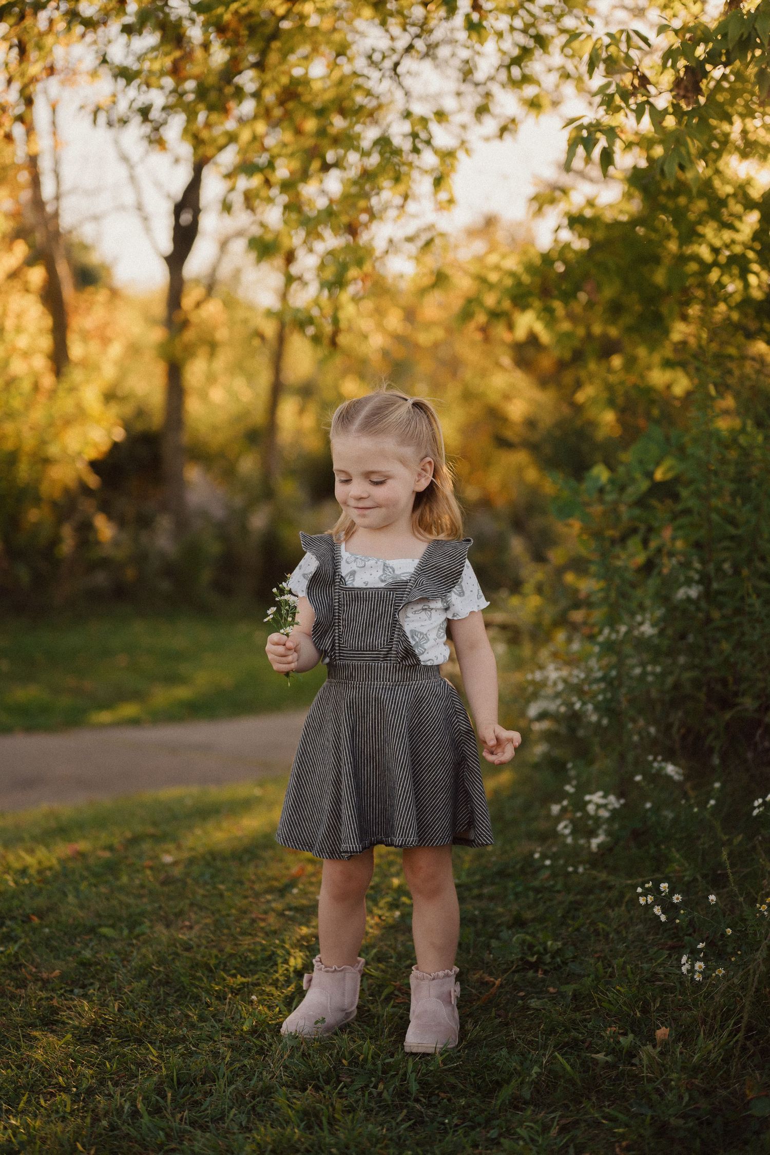A small child in a gray dress stands among wildflowers during golden hour in a natural outdoor setting.