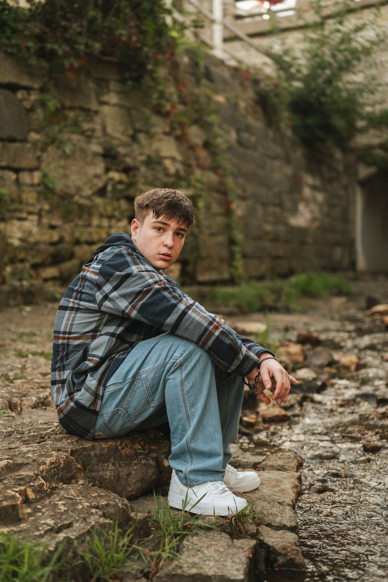 A person in a plaid shirt and jeans sits on stone ruins in a moody outdoor setting.