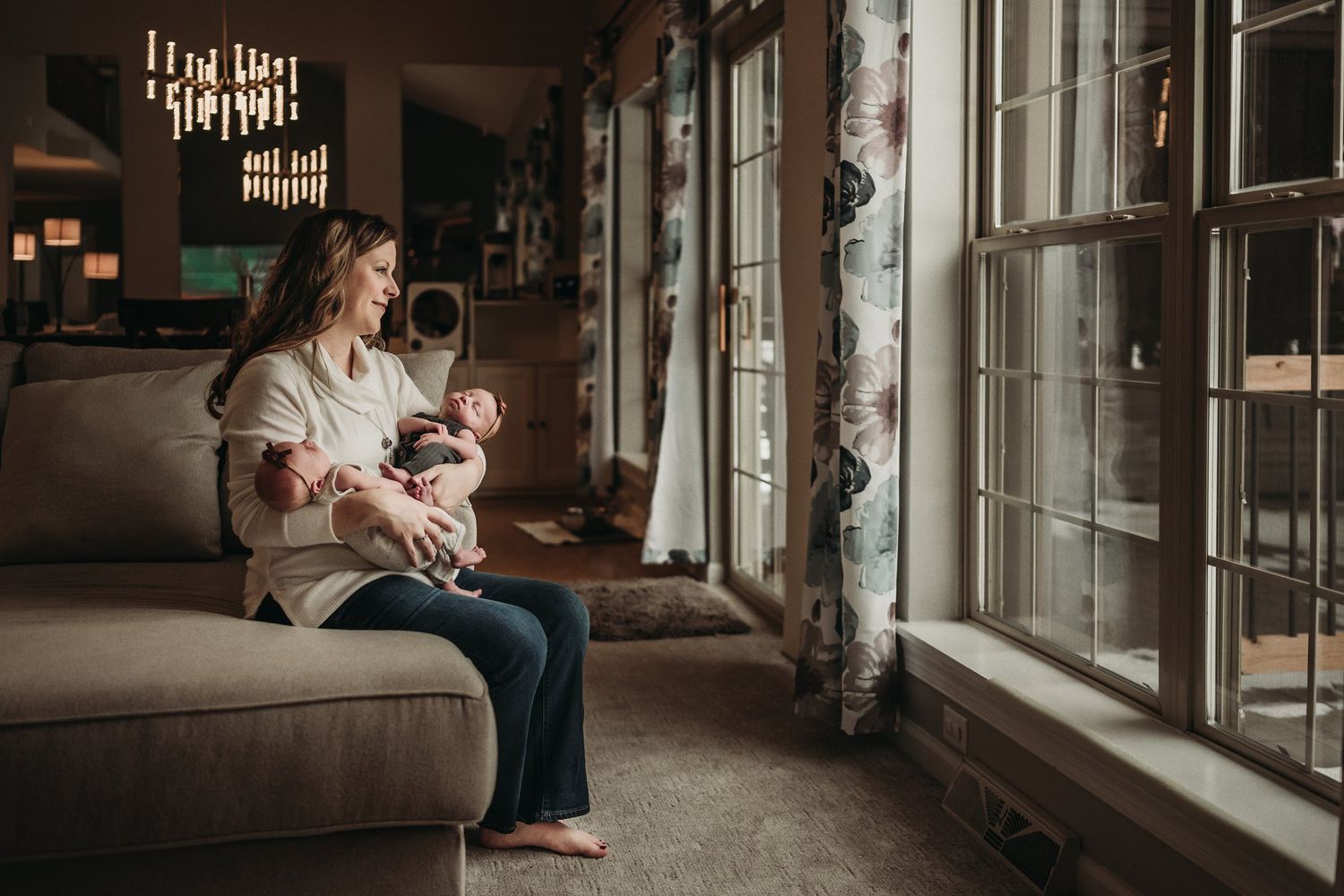 Parent and baby share quiet moment on brown sofa near large windows with natural light streaming in.