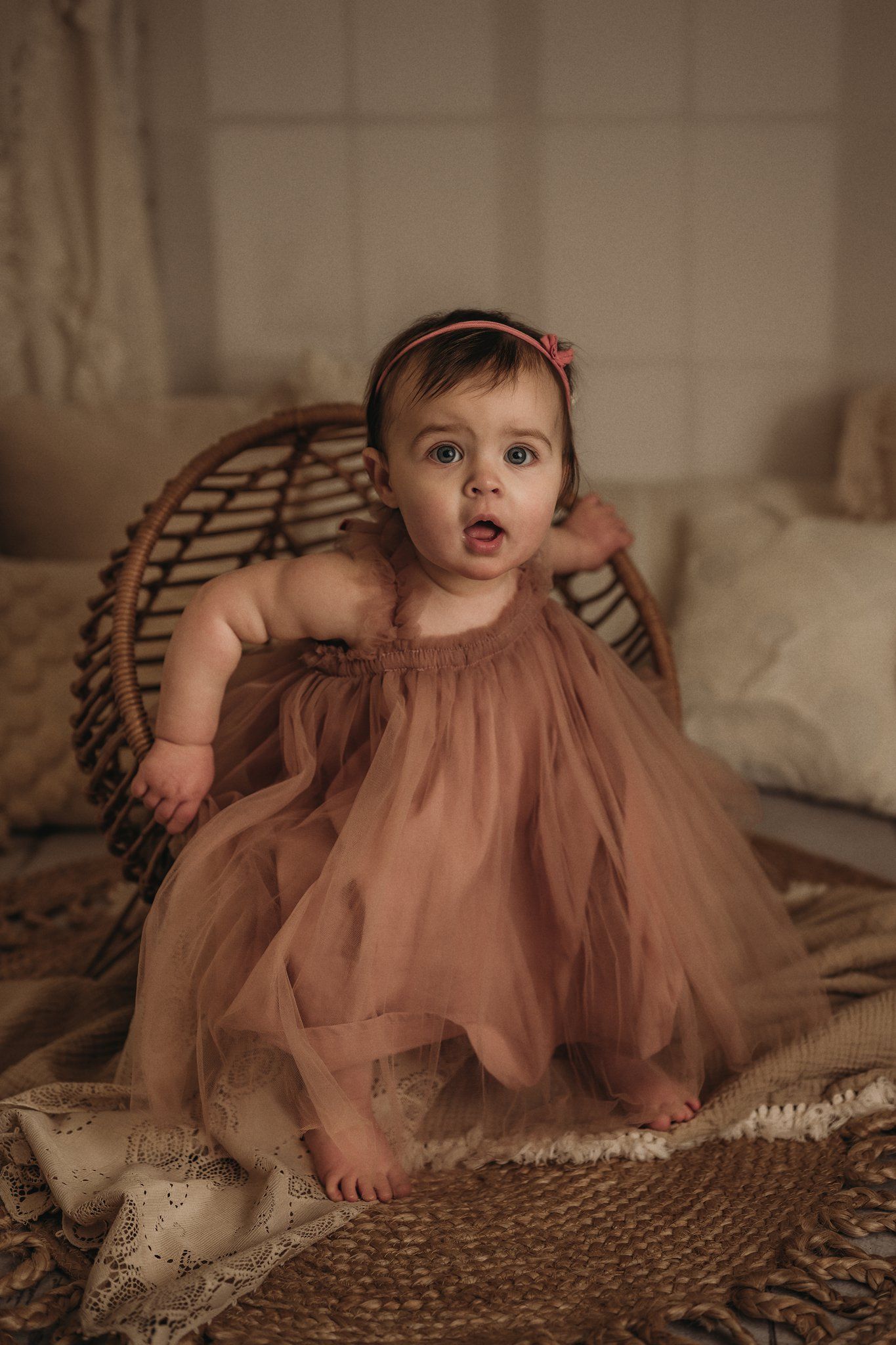Baby in dusty rose tulle dress sits on textured rug against wicker chair.