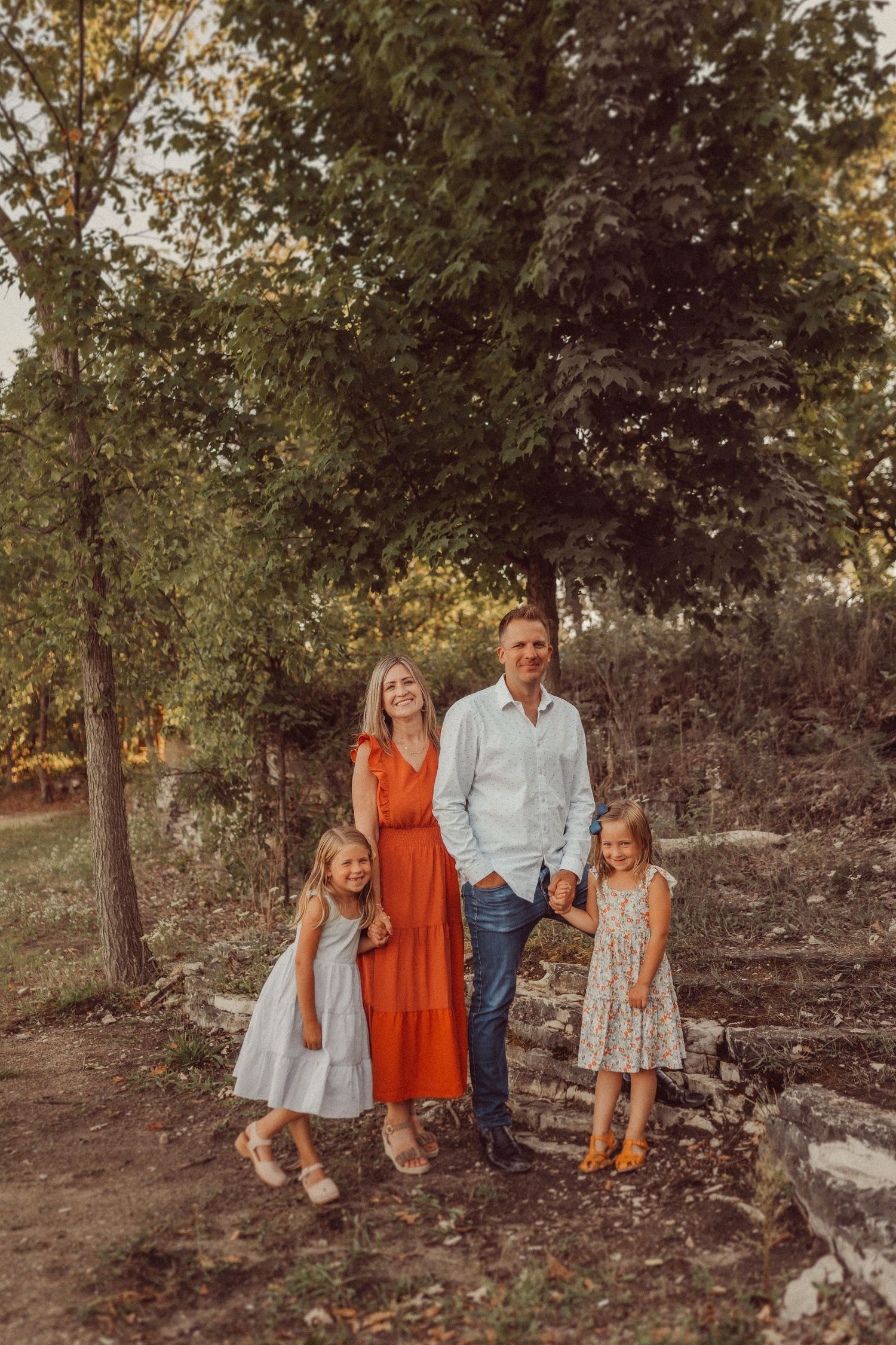 A family group poses together outdoors among trees wearing coordinated outfits in orange and blue tones.
