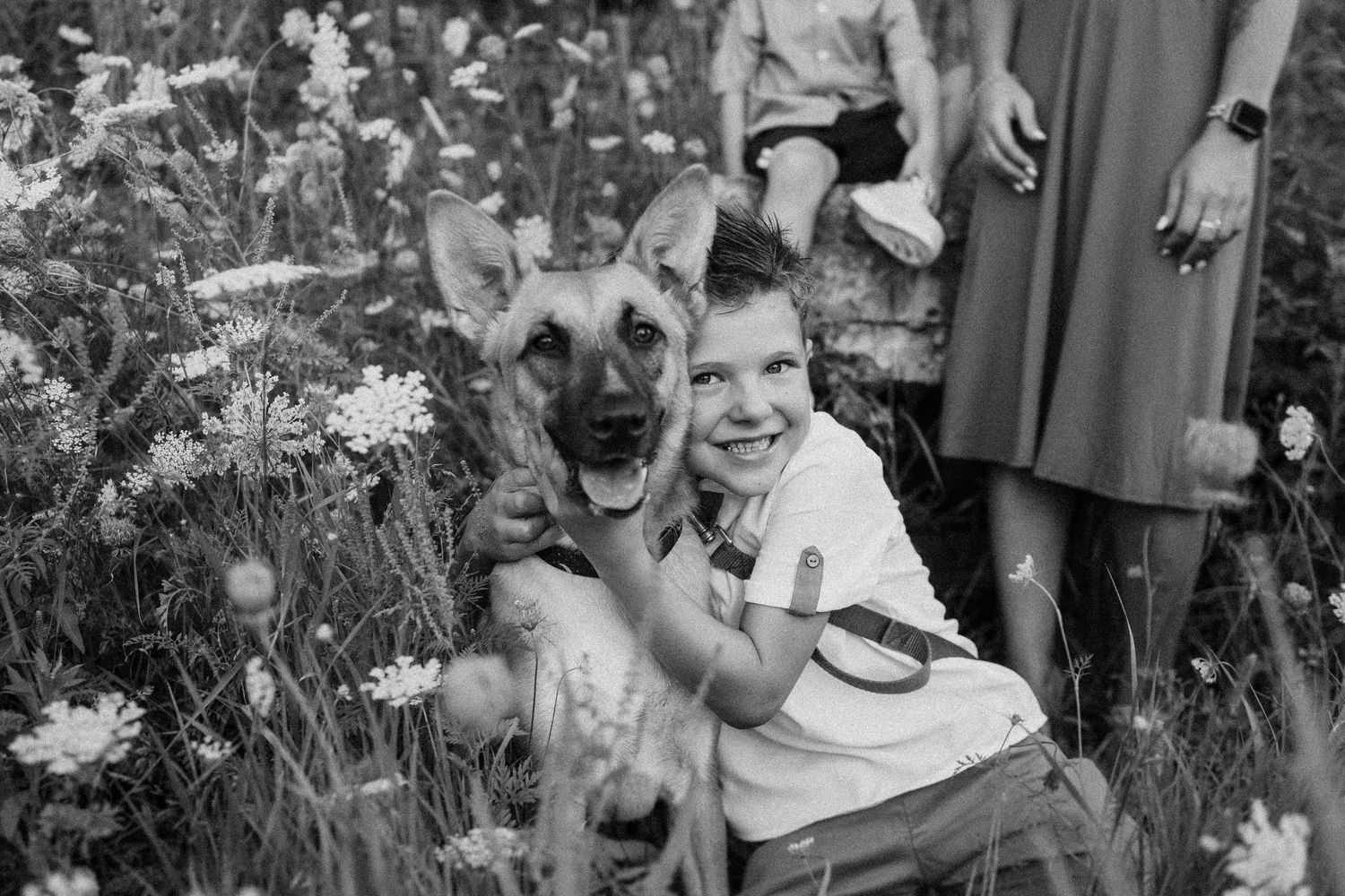 A black and white photo of a young child embracing a German Shepherd in a flower field.