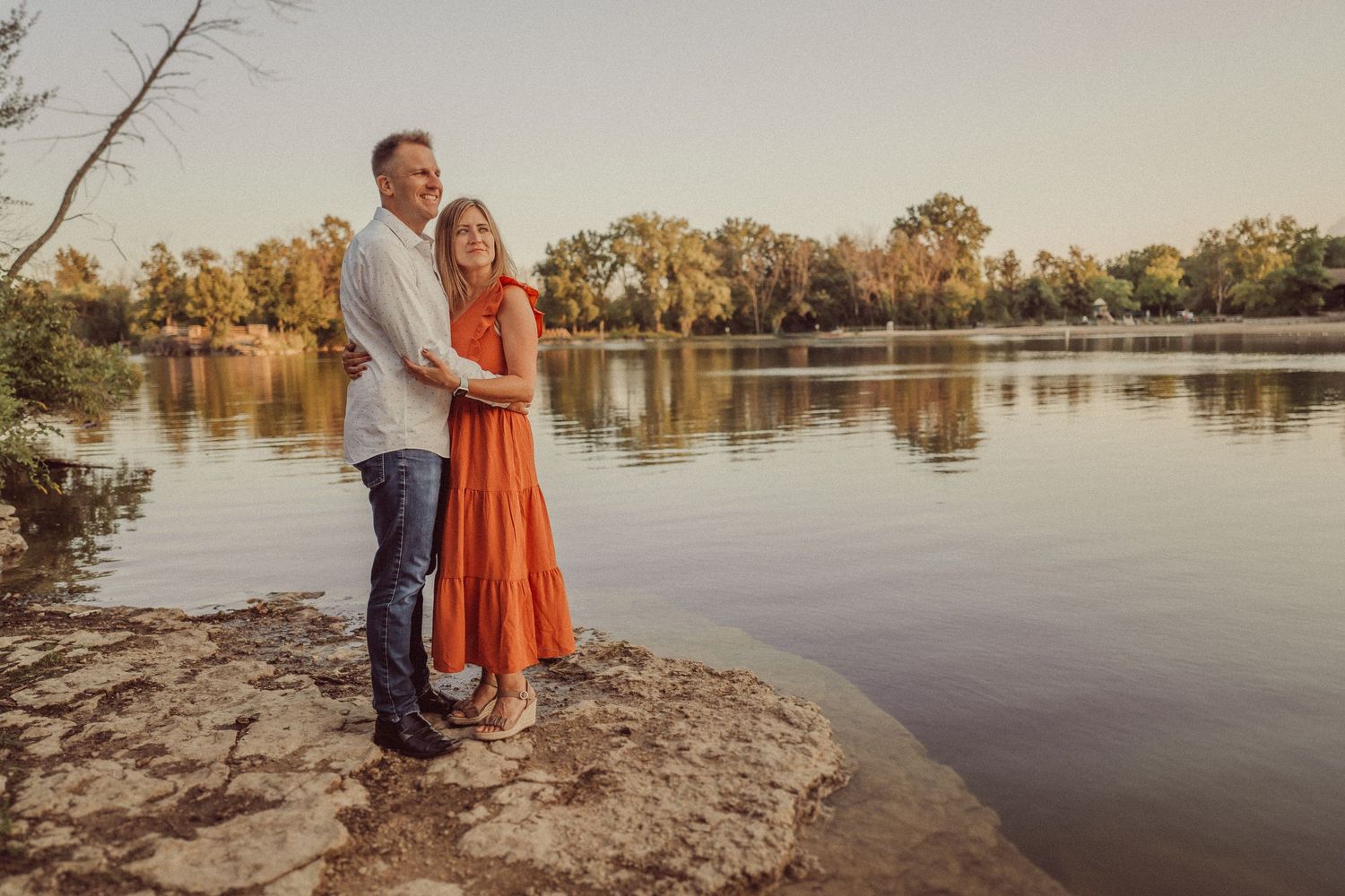 A couple stands together on a rocky shoreline beside a tranquil lake during golden hour.