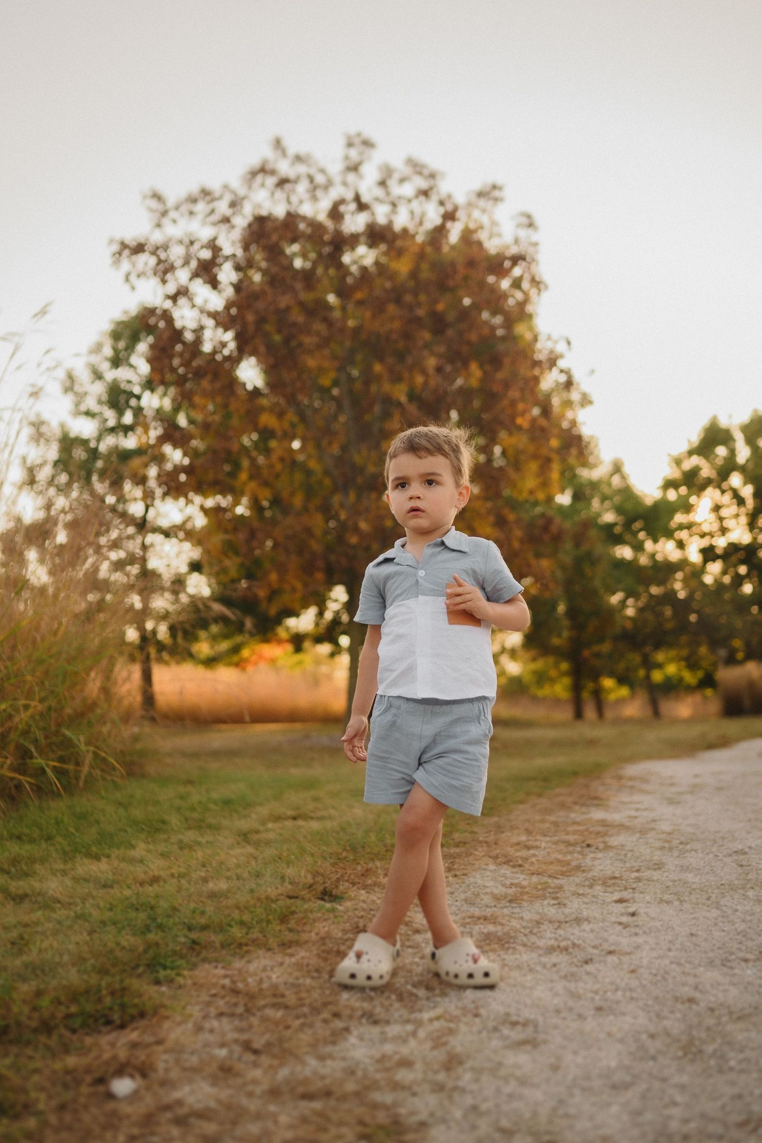 Young child in light blue outfit standing on dirt path with autumn trees in background.