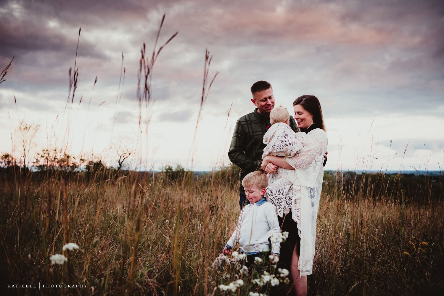 A tender family portrait against a dramatic cloudy sunset sky in a rustic field setting.