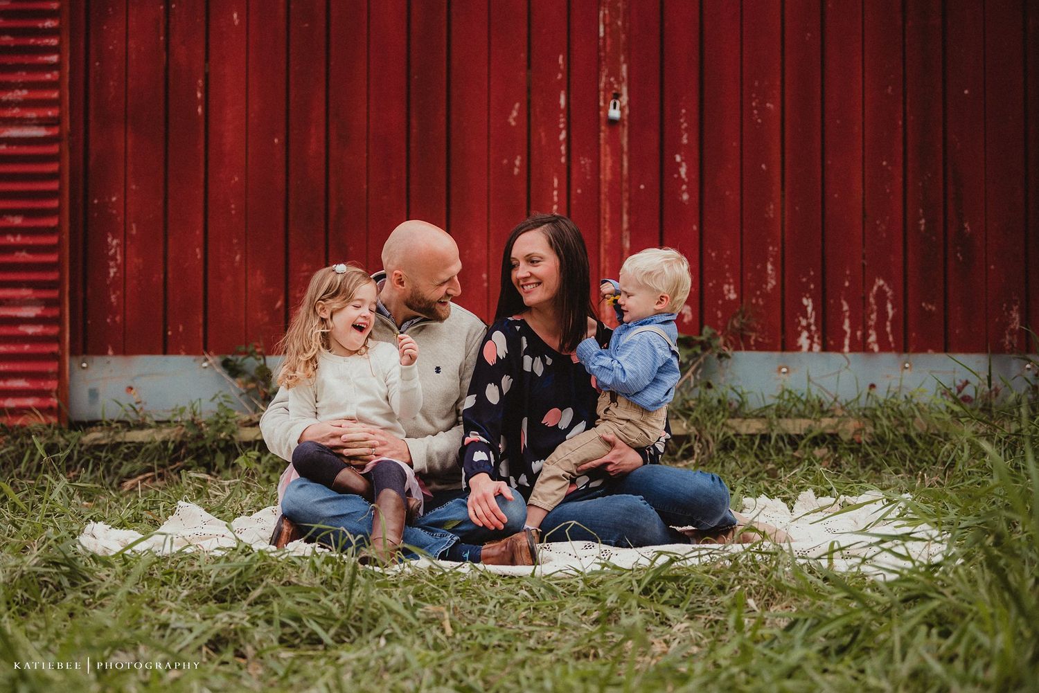 Happy family sitting together on a blanket near a rustic red barn.