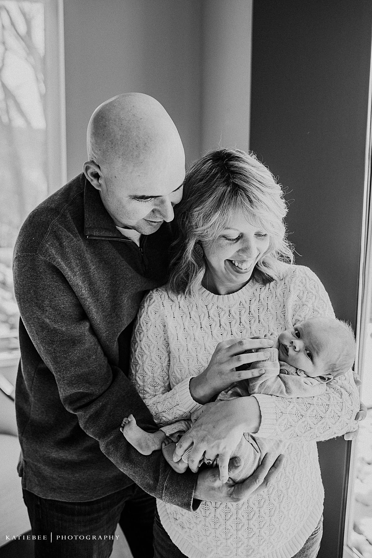 A tender black and white portrait of a couple sharing a joyful moment with their newborn baby by a window.