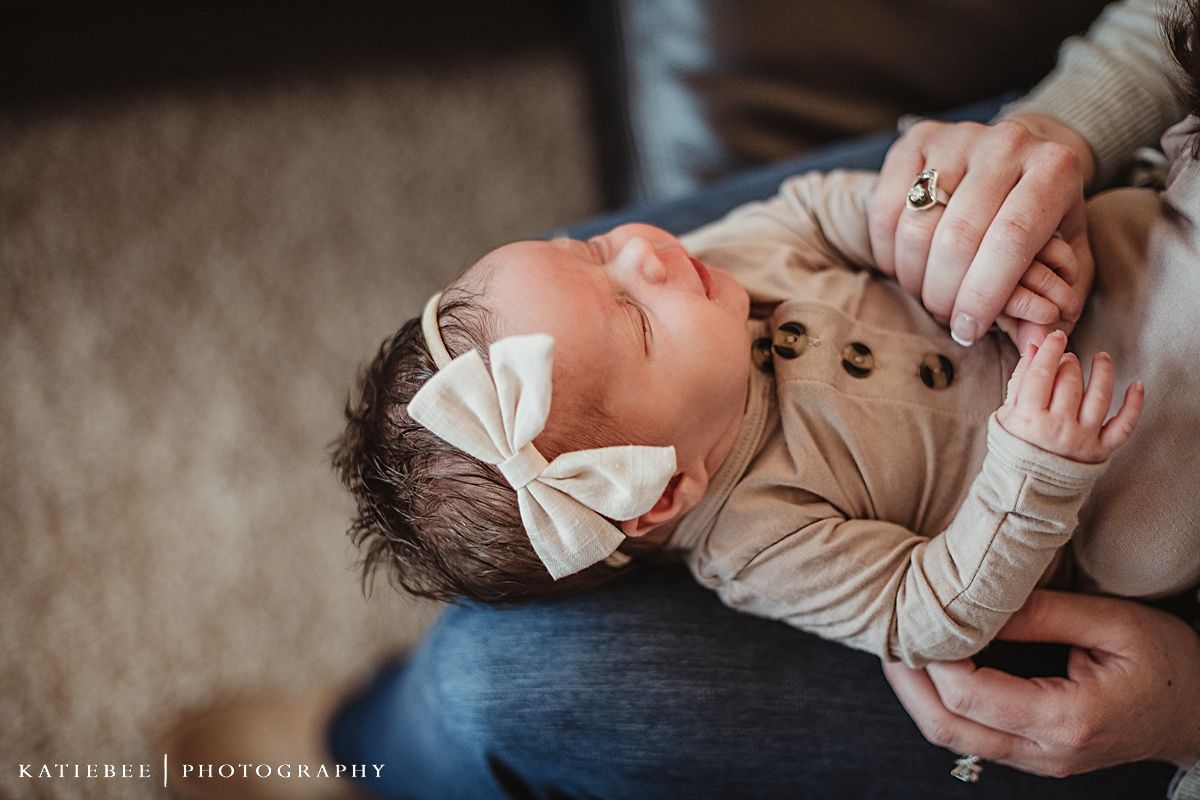 Baby wearing tan outfit with bow lying on lap during indoor lifestyle photoshoot.